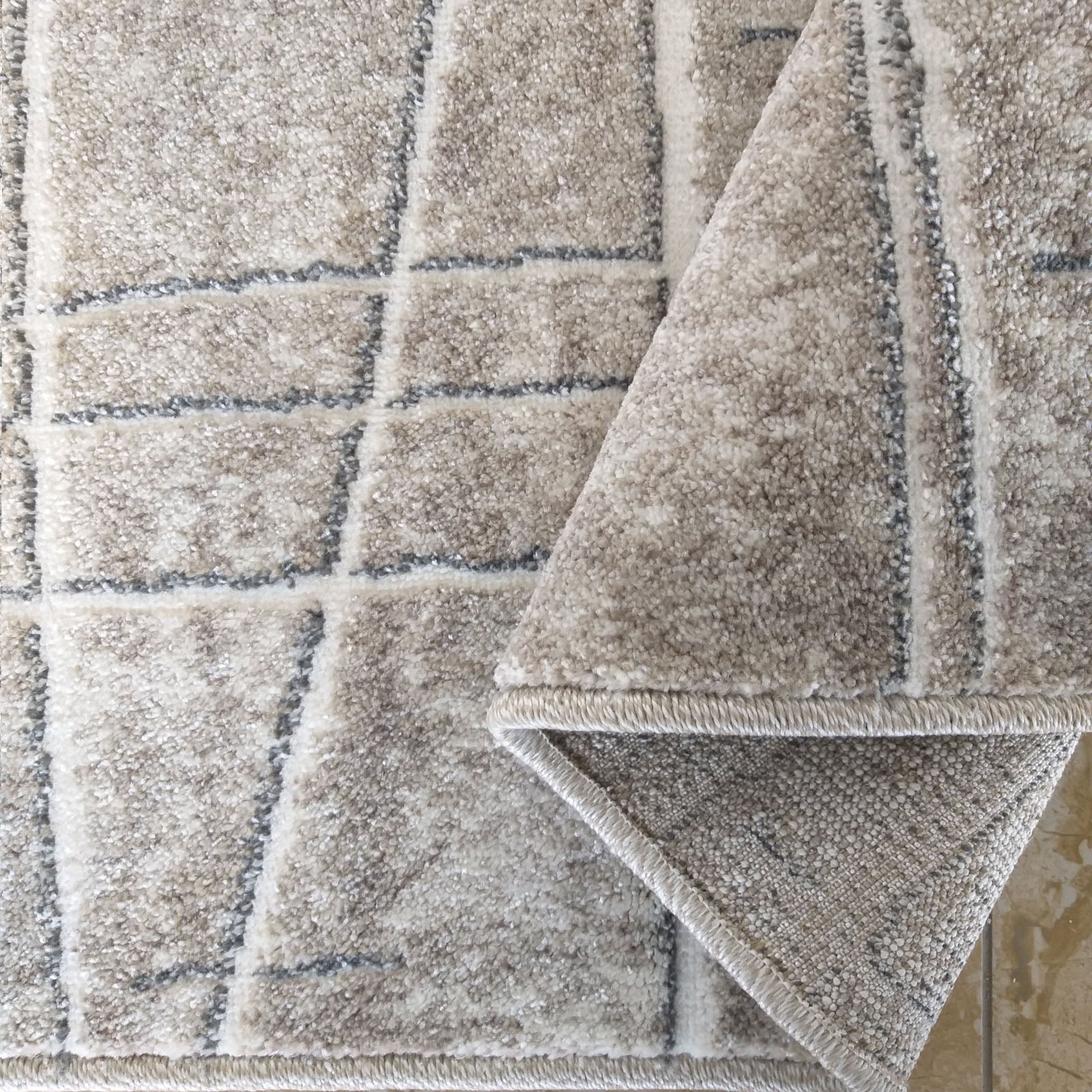 Tapis moelleux beige et crème avec un motif de carrés et lignes grises, un coin est retourné