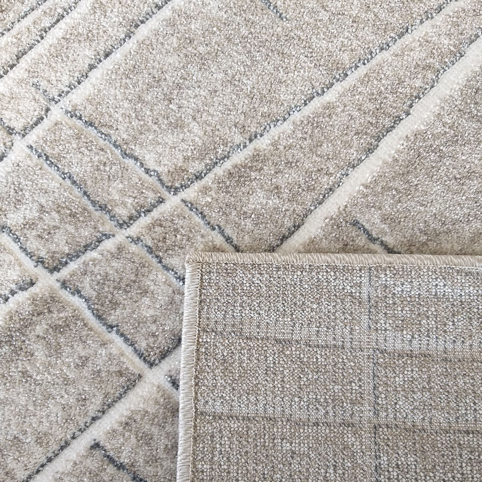 Tapis doux beige et crème avec un motif de losanges diagonaux gris. Un coin est relevé, dévoilant le revers tissé du tapis