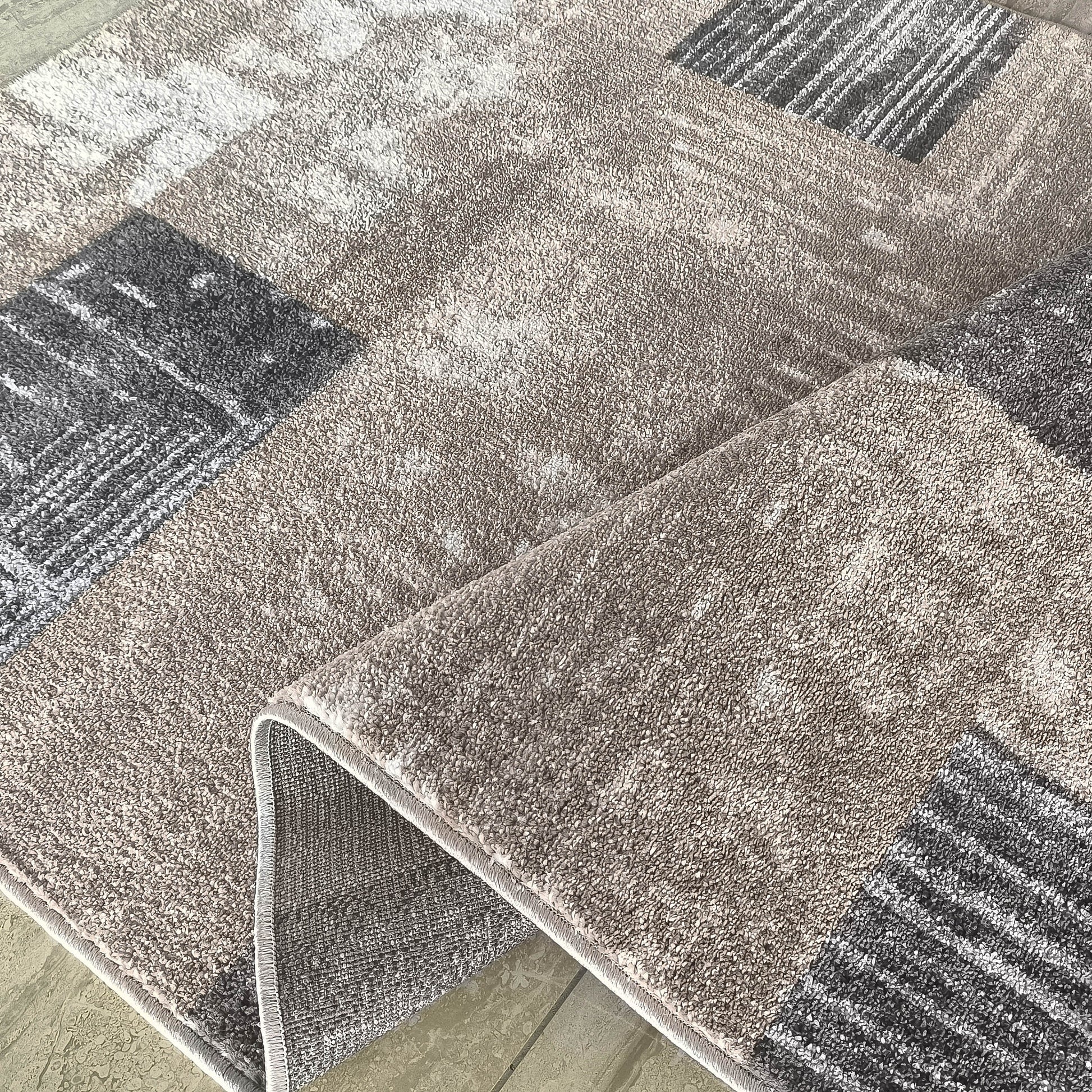 Tapis doux aux motifs abstraits et géométriques beige, gris clair et foncé, avec un coin relevé montrant le dos du tapis