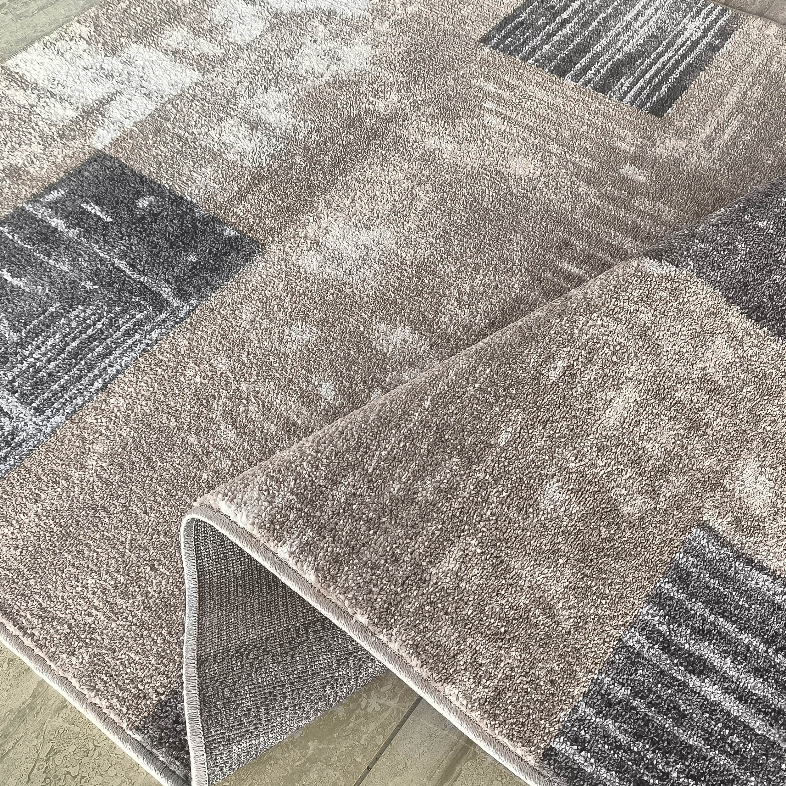 Tapis doux aux motifs abstraits et géométriques beige, gris clair et foncé, avec un coin relevé montrant le dos du tapis