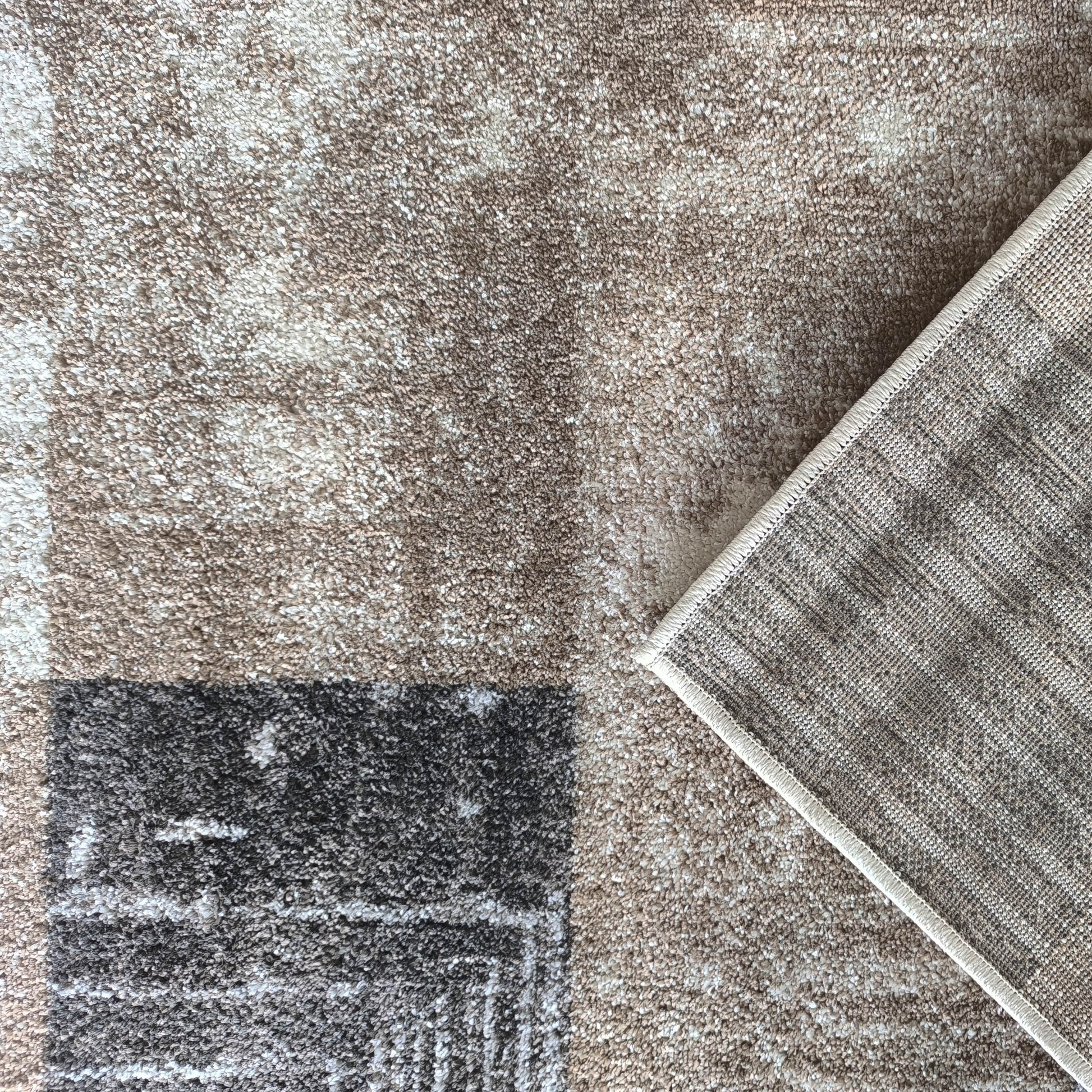 Tapis à poils doux beige, marron et gris foncé avec motifs abstraits. Un coin est plié, révélant le revers tissé du tapis