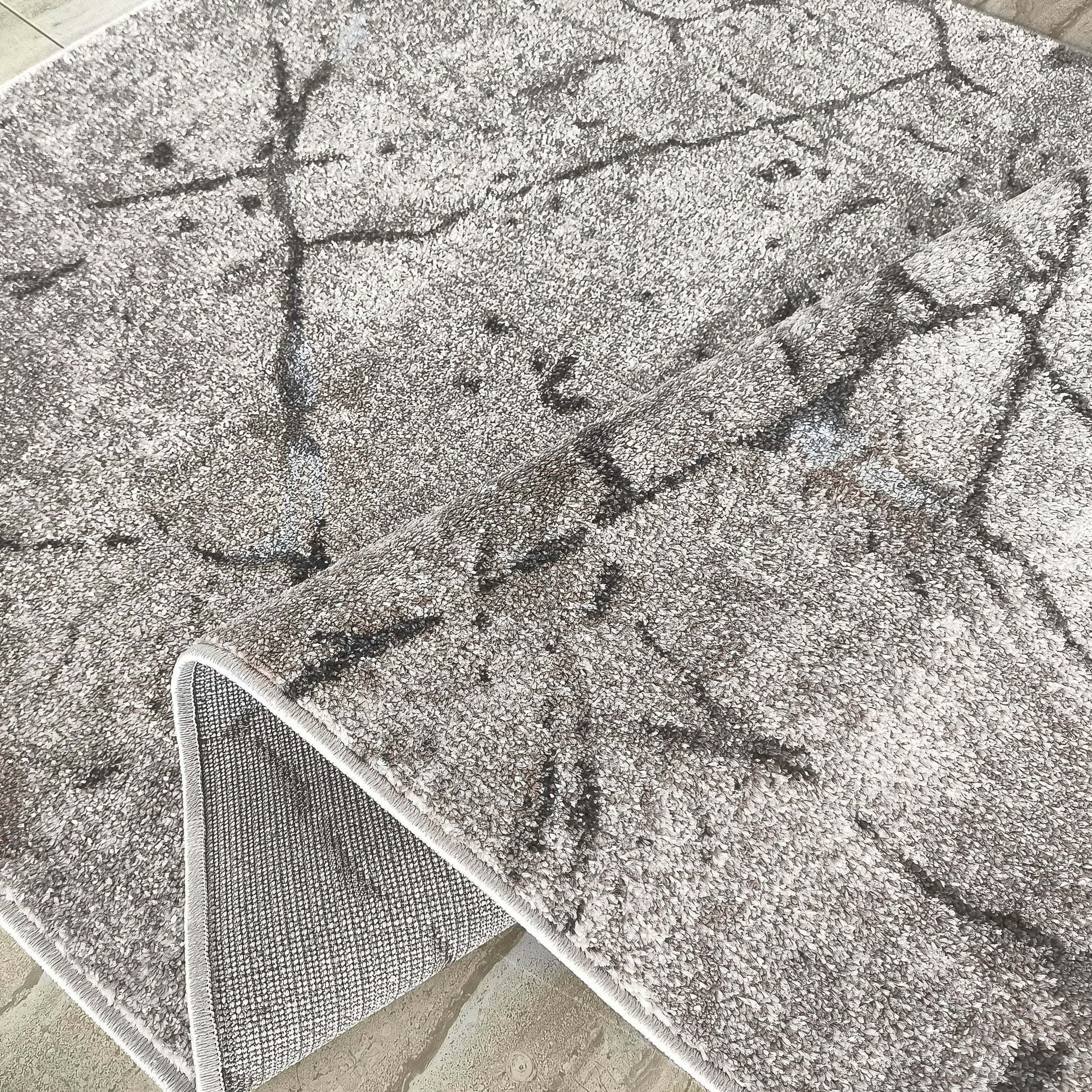 Tapis gris clair moelleux aux motifs abstraits foncés, avec un coin soulevé révélant son dos antidérapant