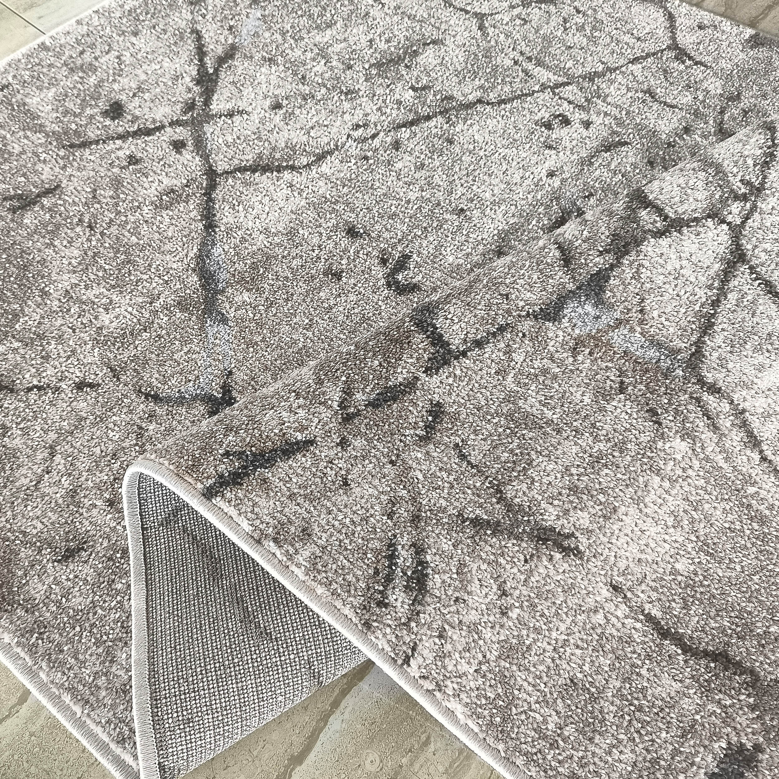 Tapis gris clair moelleux aux motifs abstraits foncés, avec un coin soulevé révélant son dos antidérapant