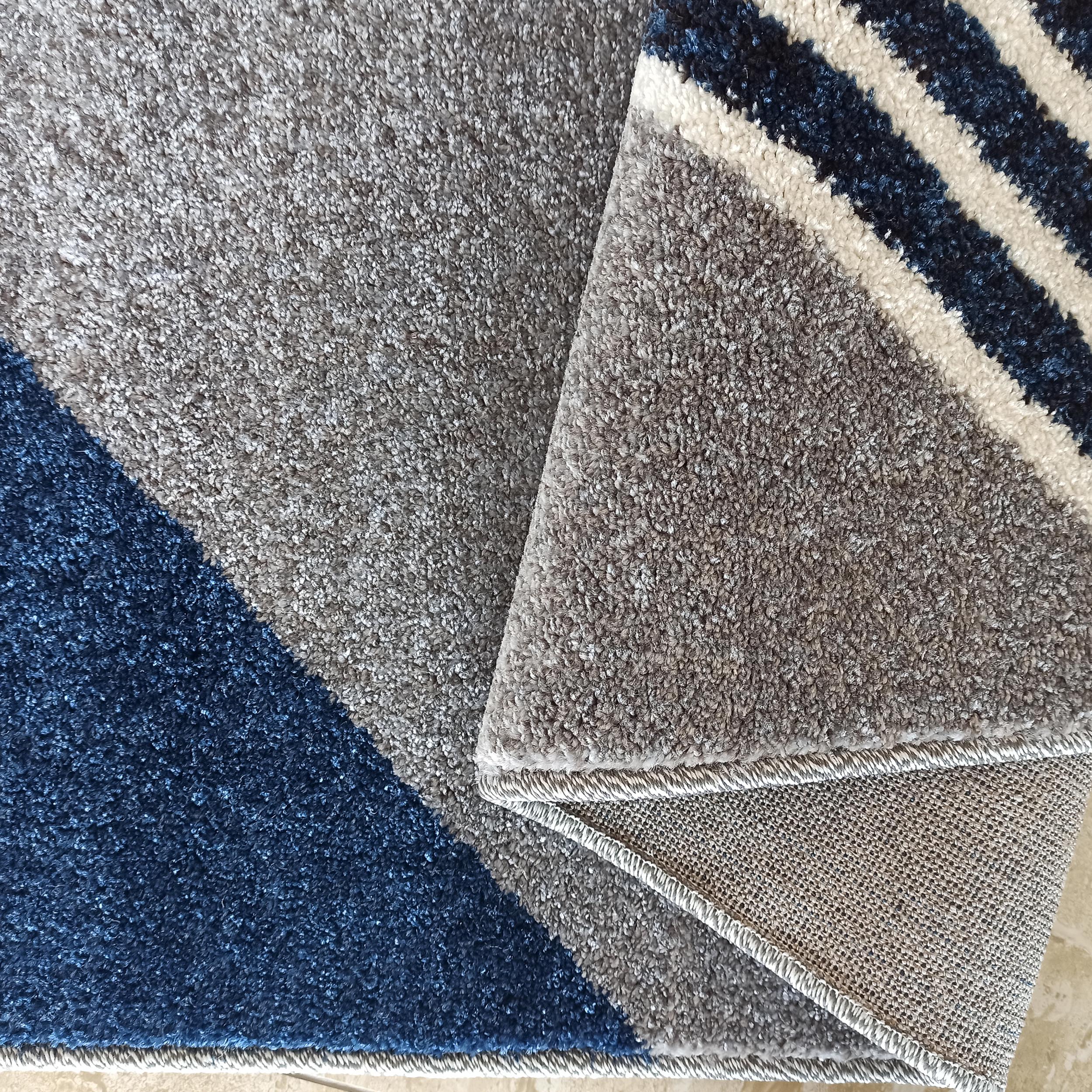 Tapis à poils longs aux motifs géométriques gris, bleu foncé et blanc, une partie est repliée laissant voir l'envers tissé