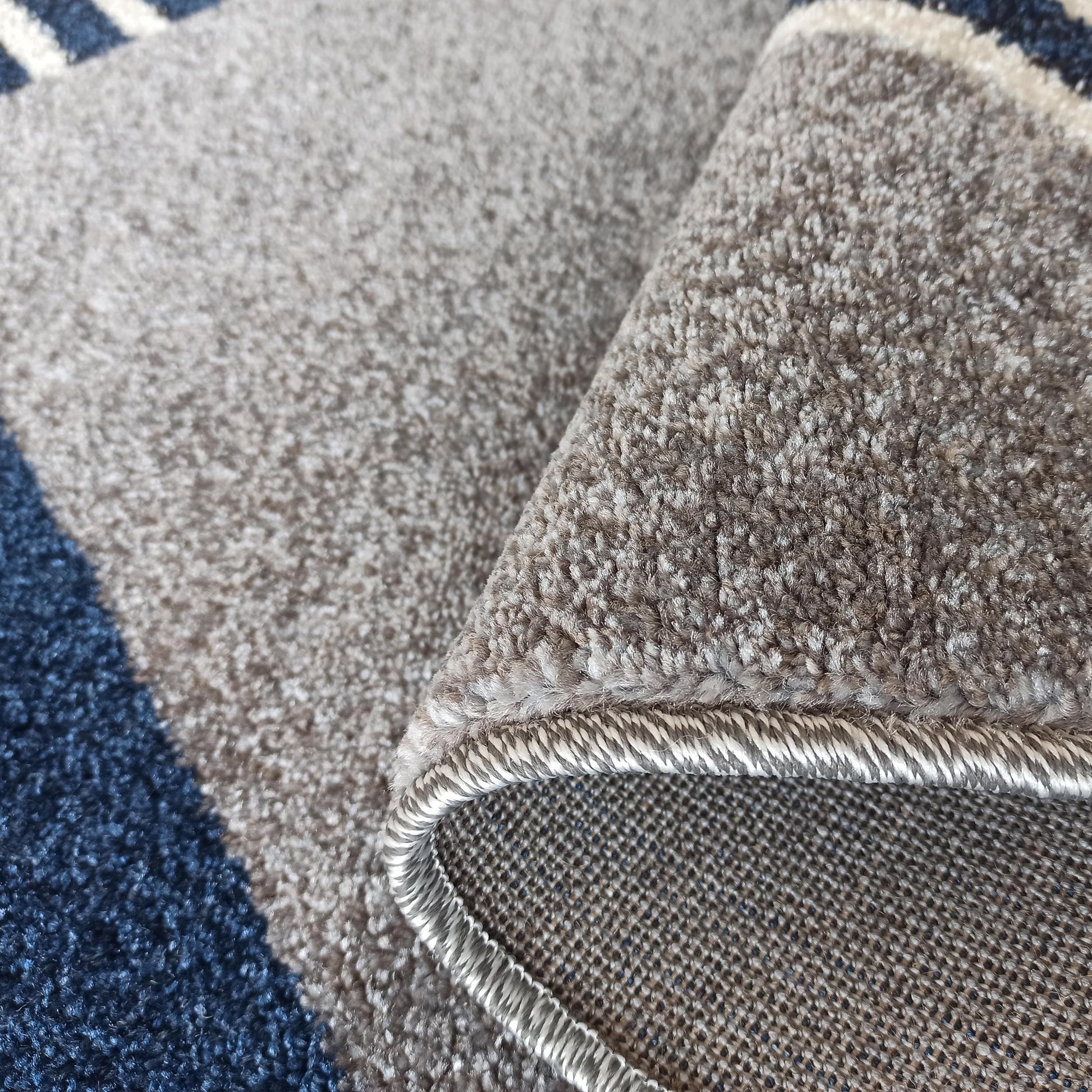 Gros plan sur un tapis doux à motifs géométriques gris, bleu foncé et blanc, en partie roulé, montrant son envers tissé robuste