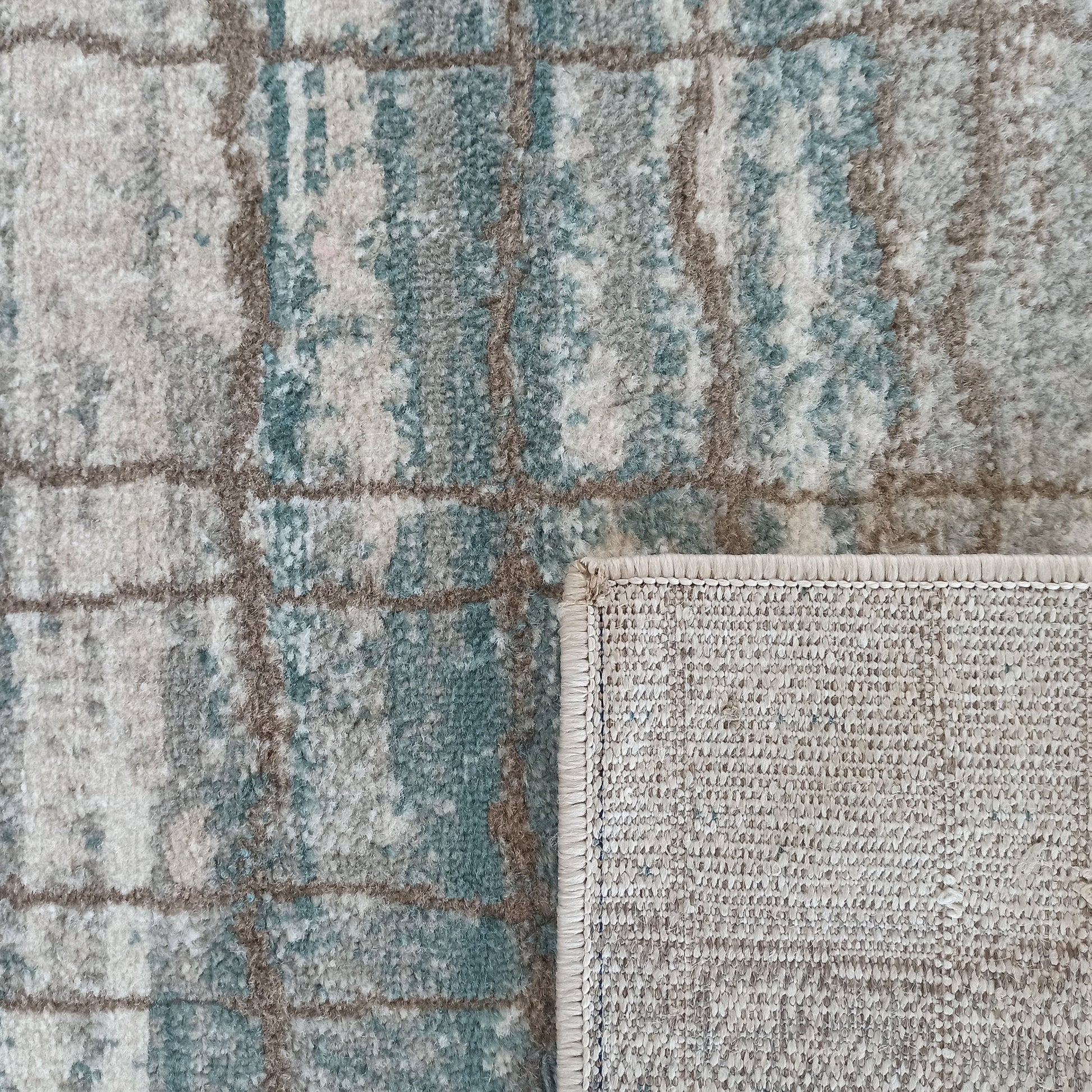 Tapis à poils longs aux motifs abstraits mêlant gris, beige clair et bleu-vert, encadrés de lignes taupe. Un coin est replié, révélant le tissage