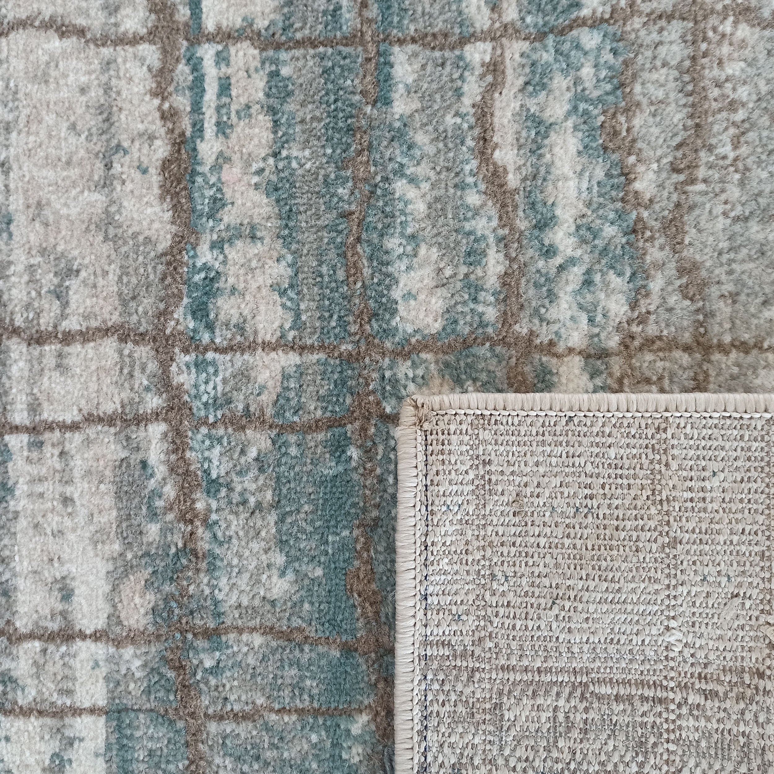 Tapis à poils longs aux motifs abstraits mêlant gris, beige clair et bleu-vert, encadrés de lignes taupe. Un coin est replié, révélant le tissage