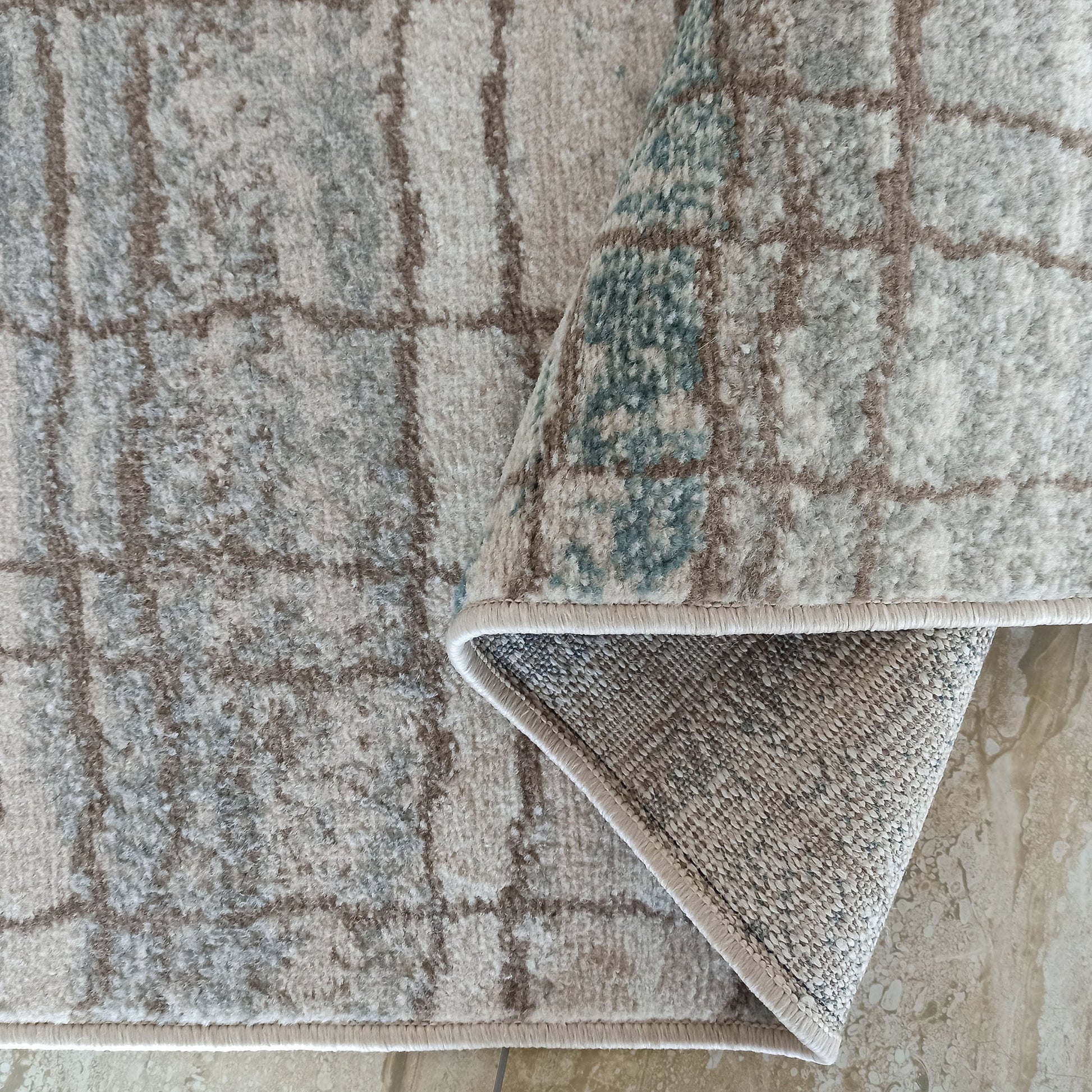 Tapis à poils courts gris, beige et bleu-vert aux motifs à carreaux irréguliers. Un coin replié révèle son envers gris tissé