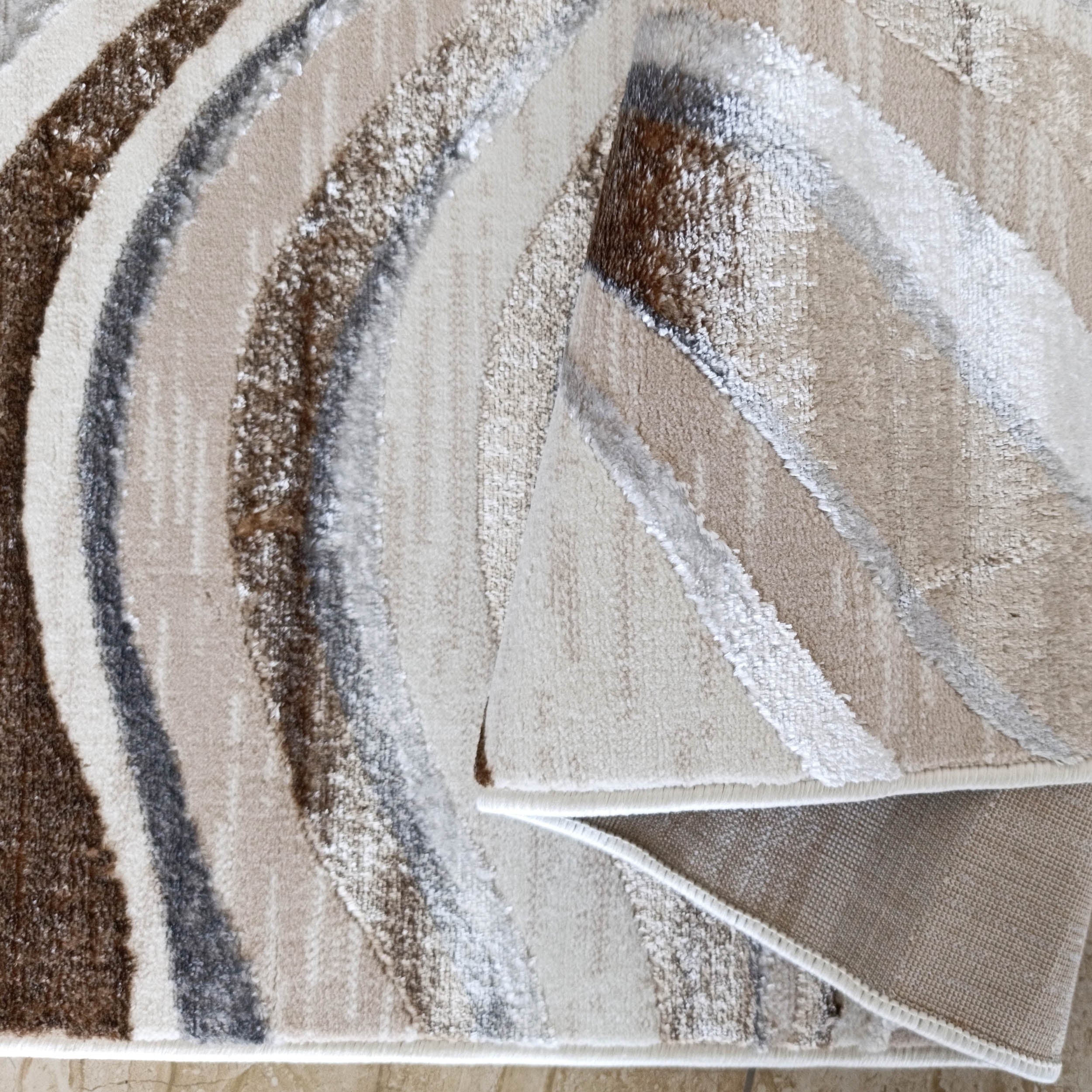 Tapis moelleux à bandes ondulées marron, beige, crème et gris dont une partie repliée révèle son envers textile clair