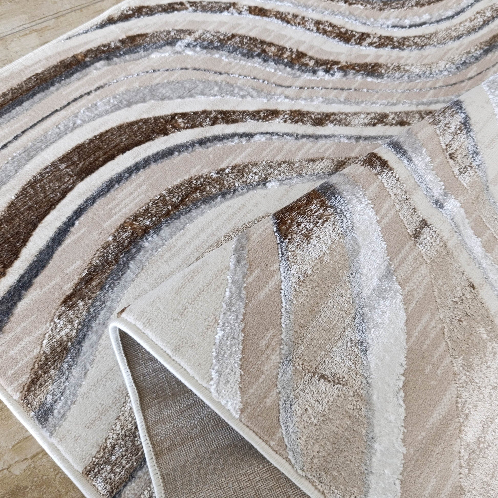 Tapis texturé aux motifs abstraits de vagues marron, grises, beiges et blanc crème, partiellement replié sur un sol carrelé