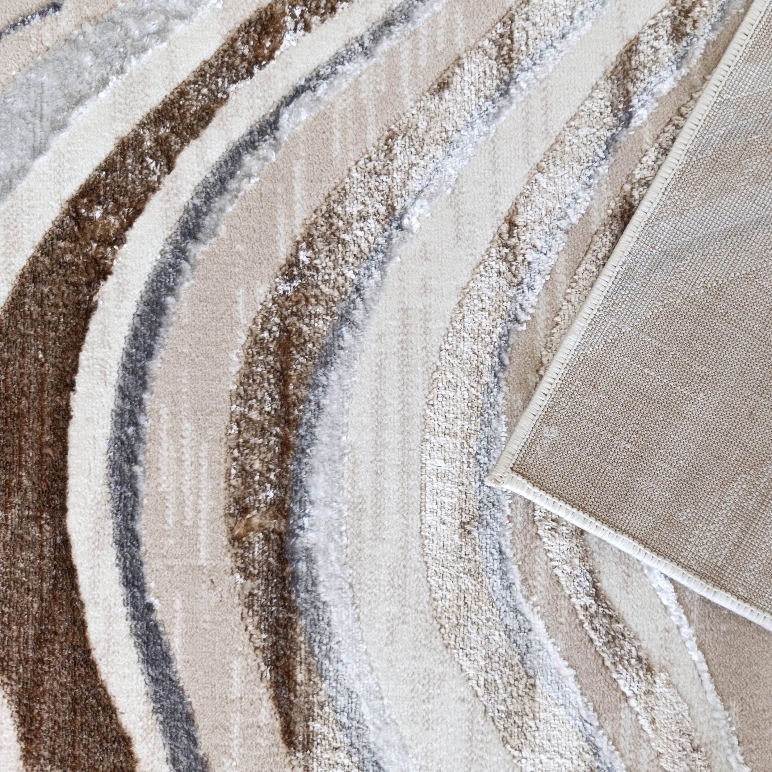 Un tapis doux aux motifs abstraits ondulés beiges, marrons, gris et blancs. Un coin est relevé, montrant son dessous tissé
