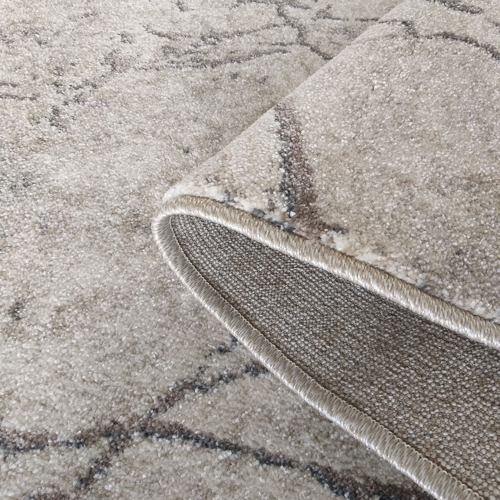Tapis moelleux aux tons clairs avec motifs abstraits gris foncé, dont un bord roulé révèle l'envers