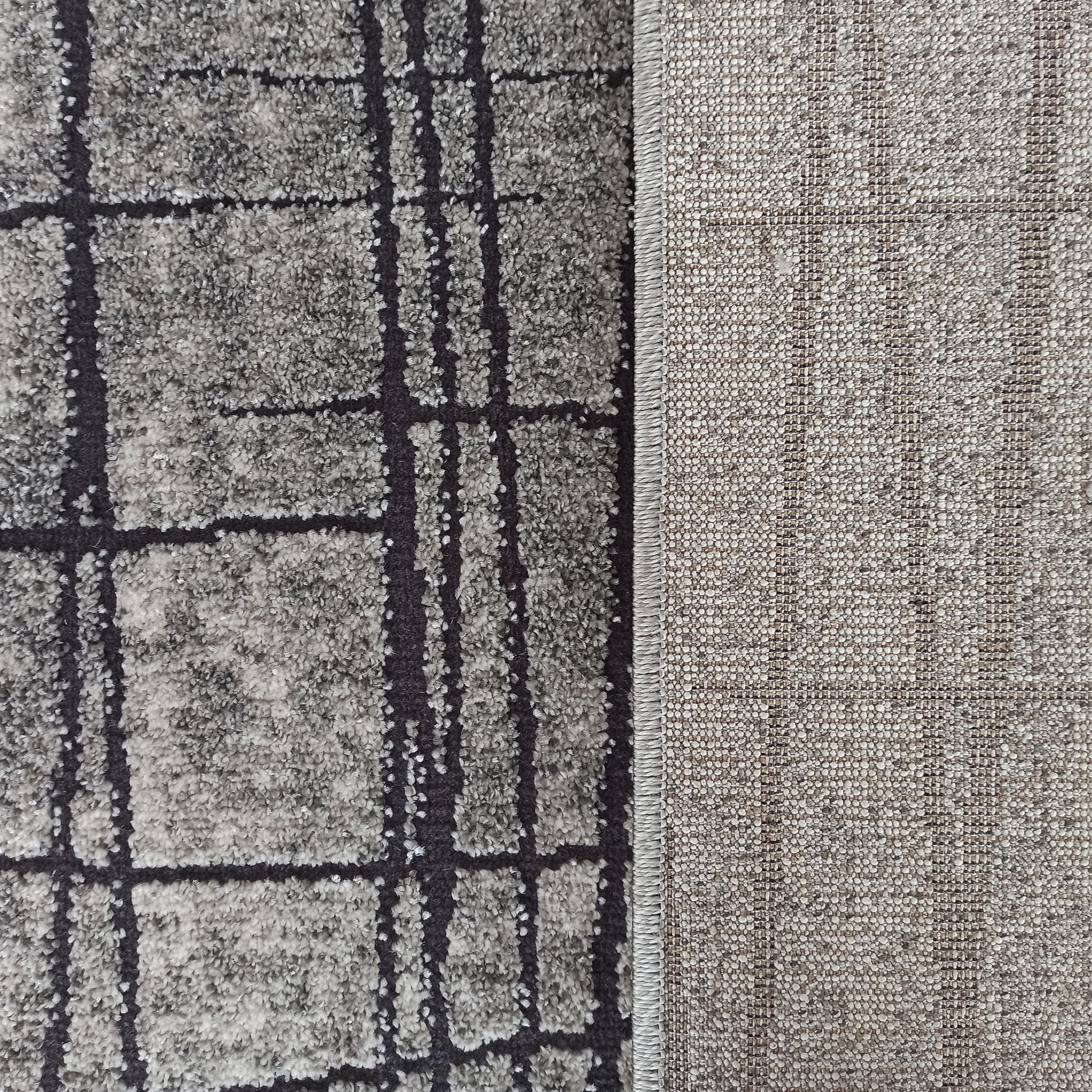 Tapis texturé gris chiné à motifs géométriques noirs, avec son revers en tissu tissé beige et bordure cousue