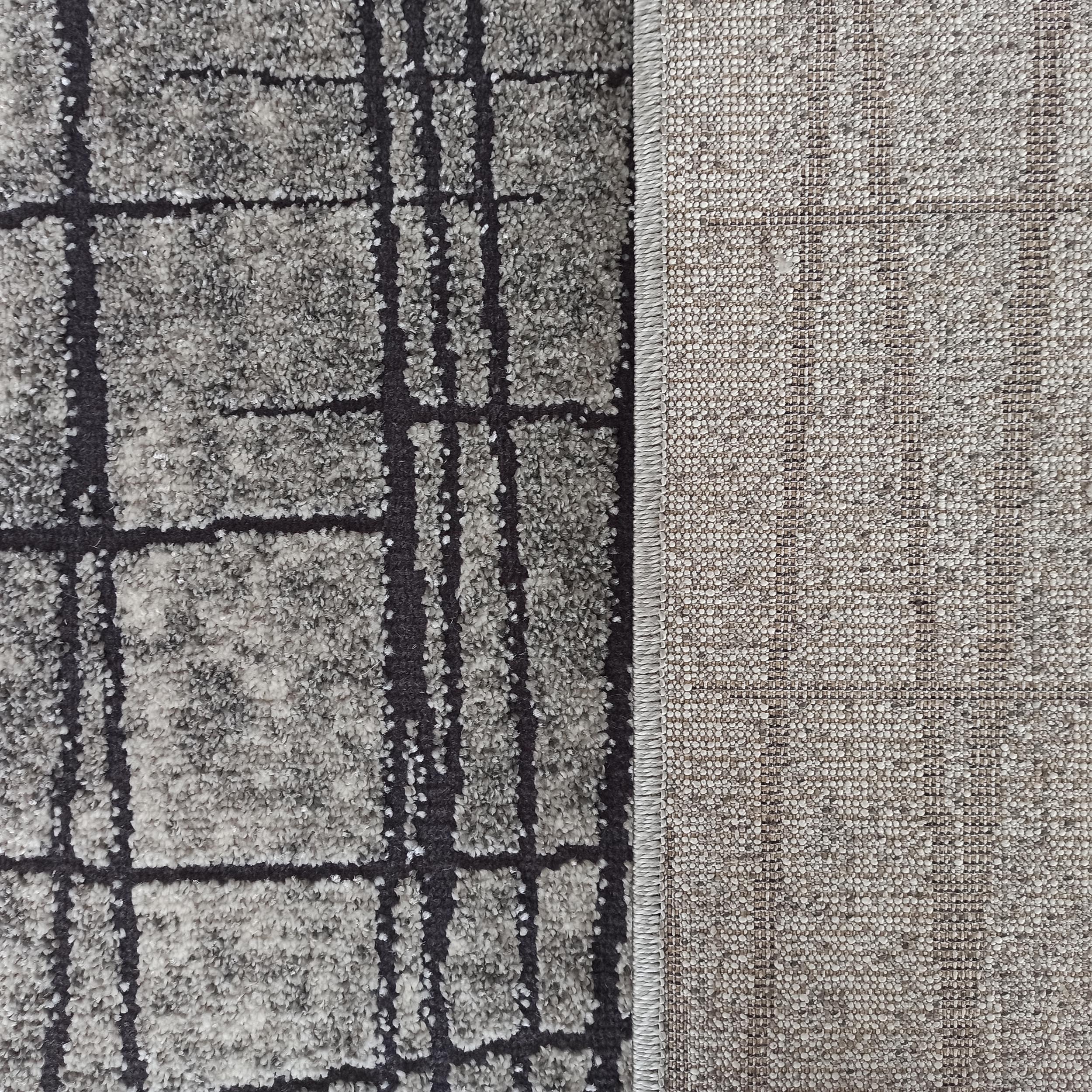 Tapis texturé gris chiné à motifs géométriques noirs, avec son revers en tissu tissé beige et bordure cousue