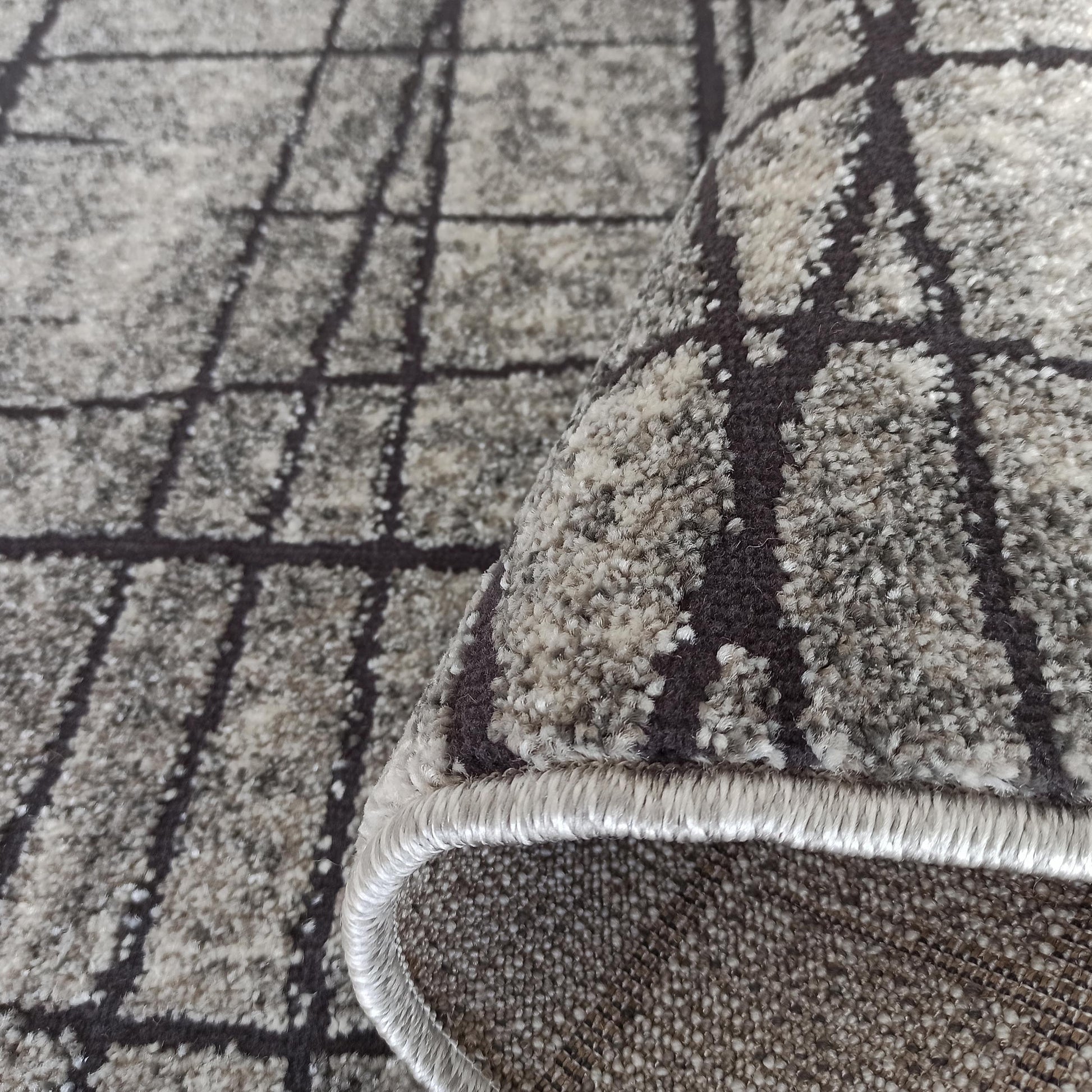 Tapis texturé gris et beige clair, motif quadrillé de lignes foncées, avec un coin roulé montrant sa bordure ourlée