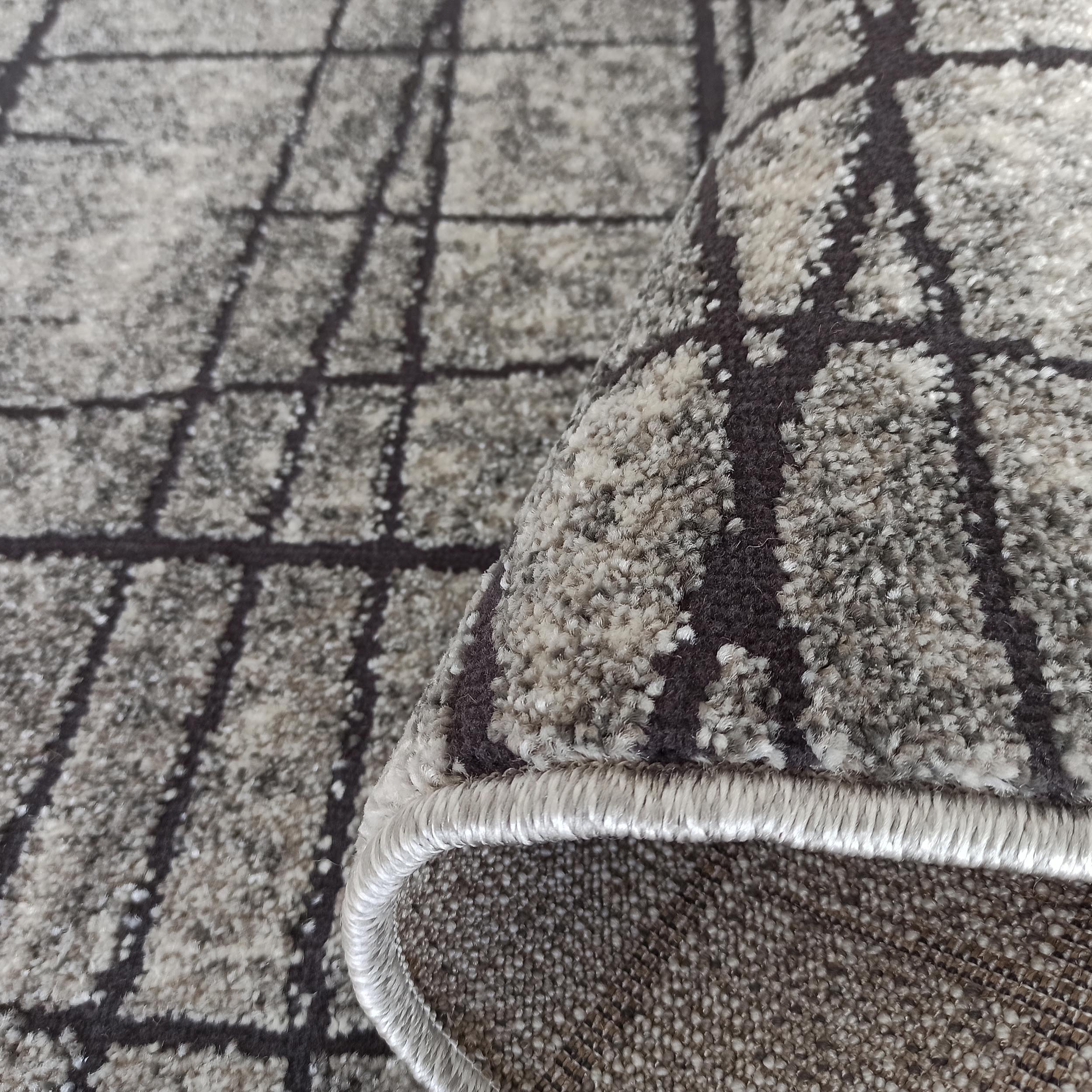 Tapis texturé gris et beige clair, motif quadrillé de lignes foncées, avec un coin roulé montrant sa bordure ourlée
