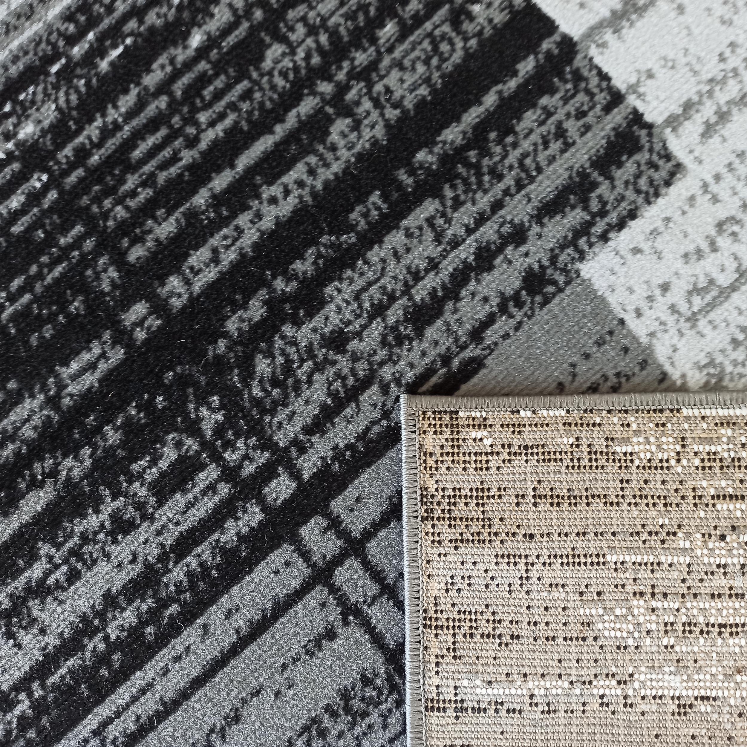 Vue rapprochée d'un tapis aux motifs abstraits noirs, gris foncés et blancs, avec l'envers beige à points noirs visible