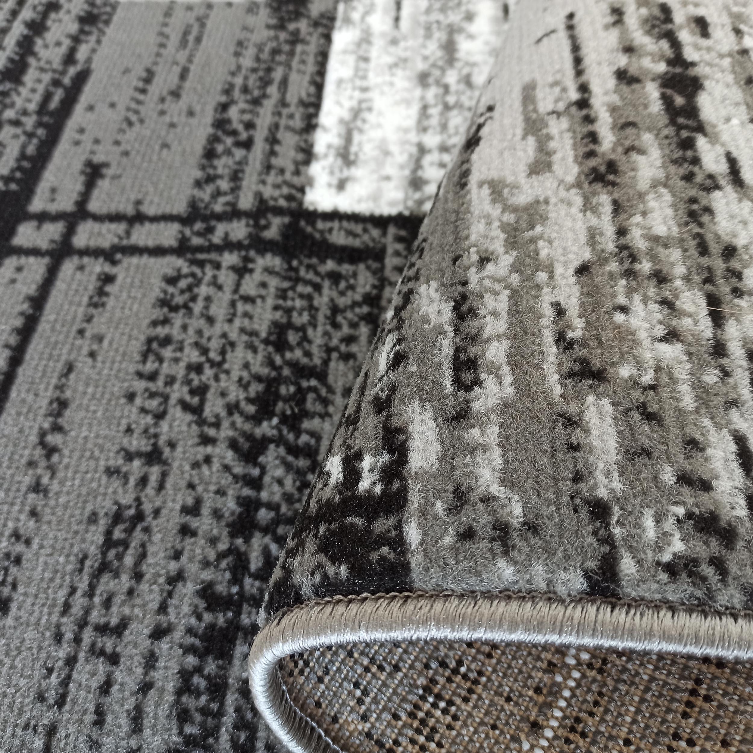 Gros plan sur un tapis aux motifs abstraits gris, noirs et blancs, dont un bord est roulé