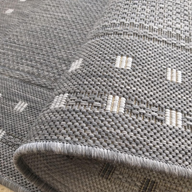 Tapis tissé gris chiné avec des motifs en petits rectangles clairs et beiges, une partie du tapis est roulée