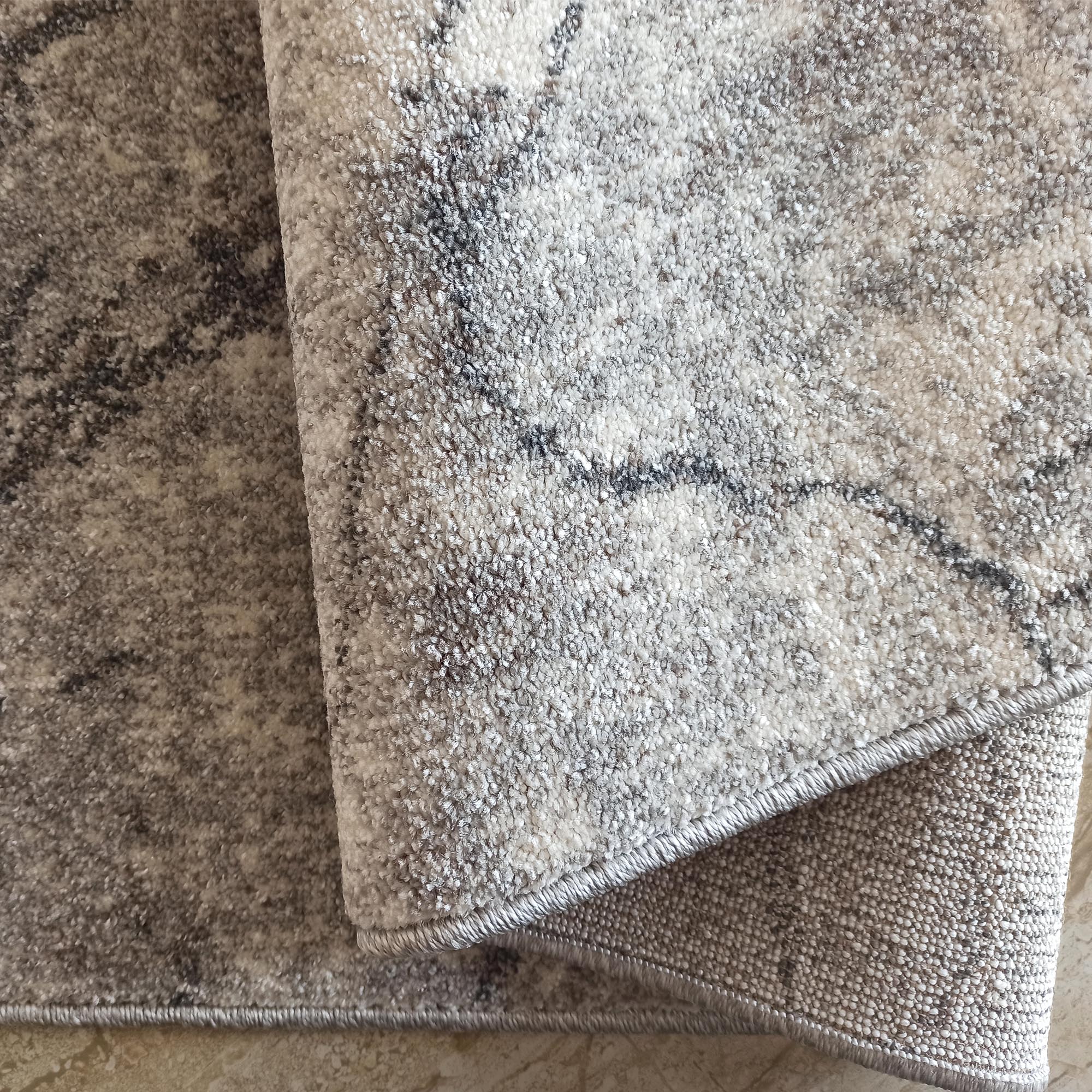 Gros plan sur un tapis texturé gris et blanc aux motifs abstraits, avec un bord surpiqué visible sur le tapis enroulé