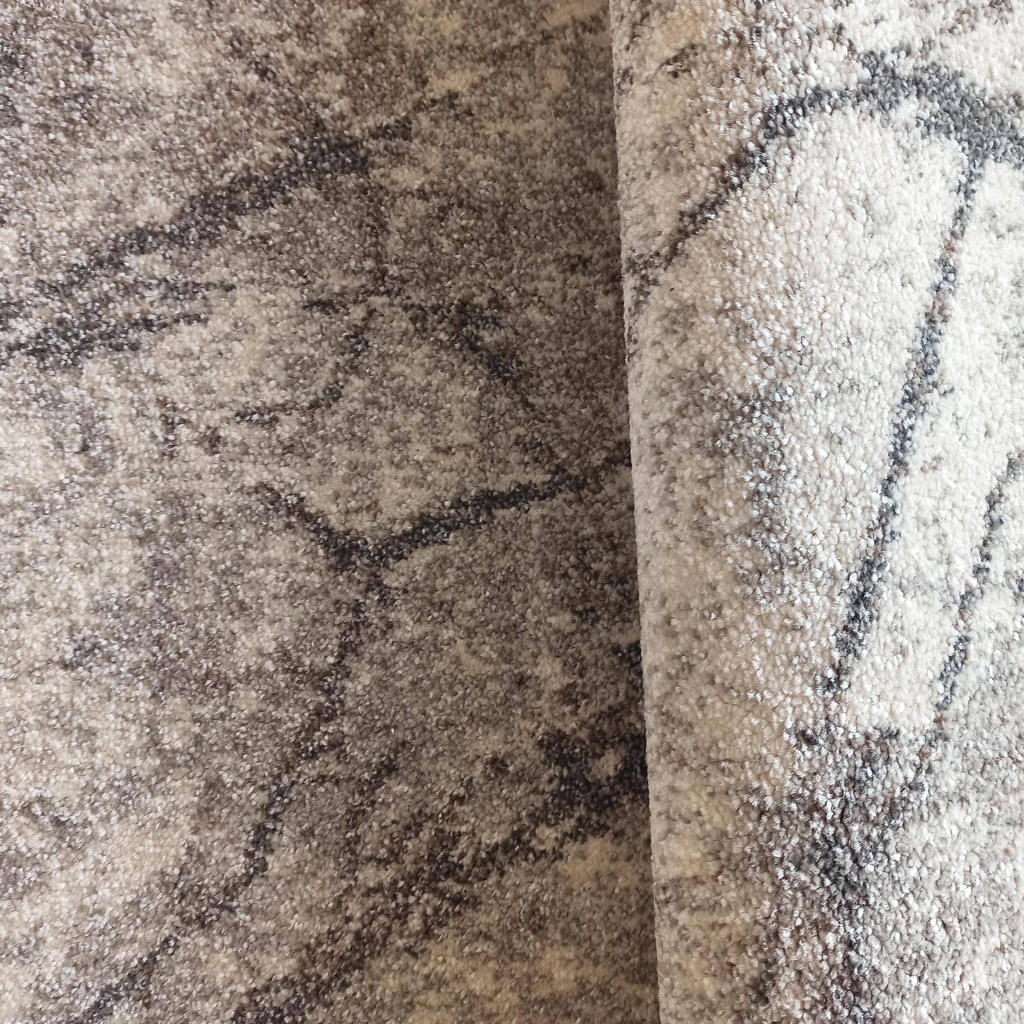 Tapis texturé aux nuances de beige clair, crème et marron. Il présente des motifs abstraits foncés et est légèrement roulé sur le côté
