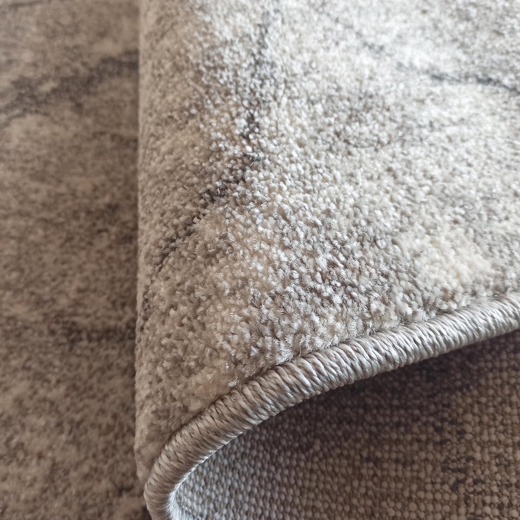 Gros plan d'un tapis doux et moelleux aux tons gris clair et beige avec des motifs abstraits foncés, montrant sa bordure