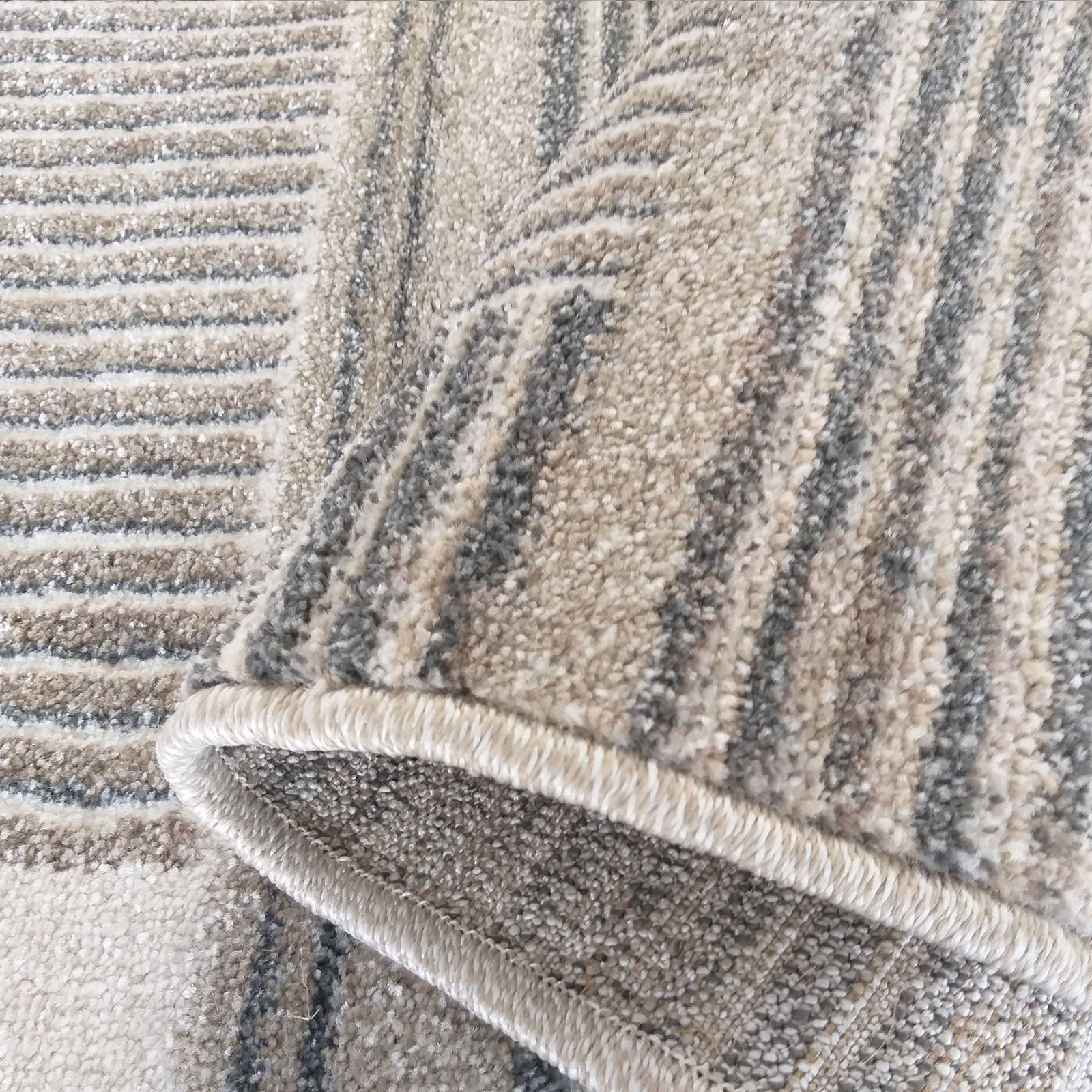 Tapis texturé aux motifs de rayures horizontales et ondulées beiges, grises et blanches, avec un liseré visible sur le côté replié