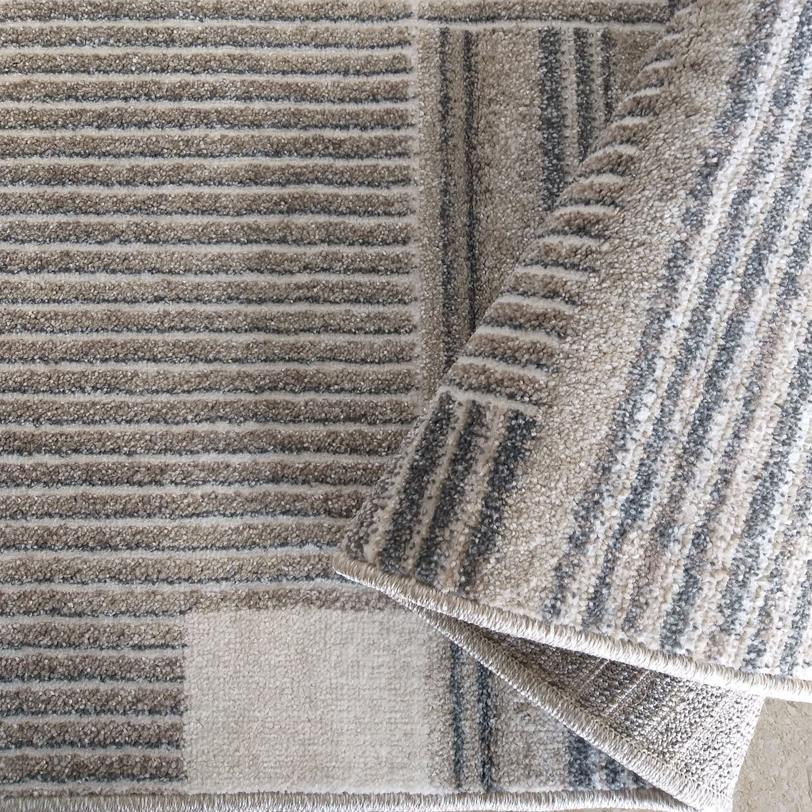 Tapis texturé beige, gris et blanc cassé avec des motifs géométriques et des rayures, dont une section est repliée