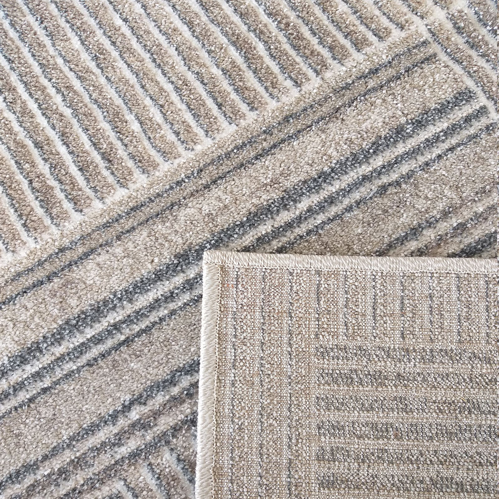 Tapis moelleux aux motifs de bandes diagonales alternant beige, gris et crème. Un coin est relevé, montrant le dessous tissé
