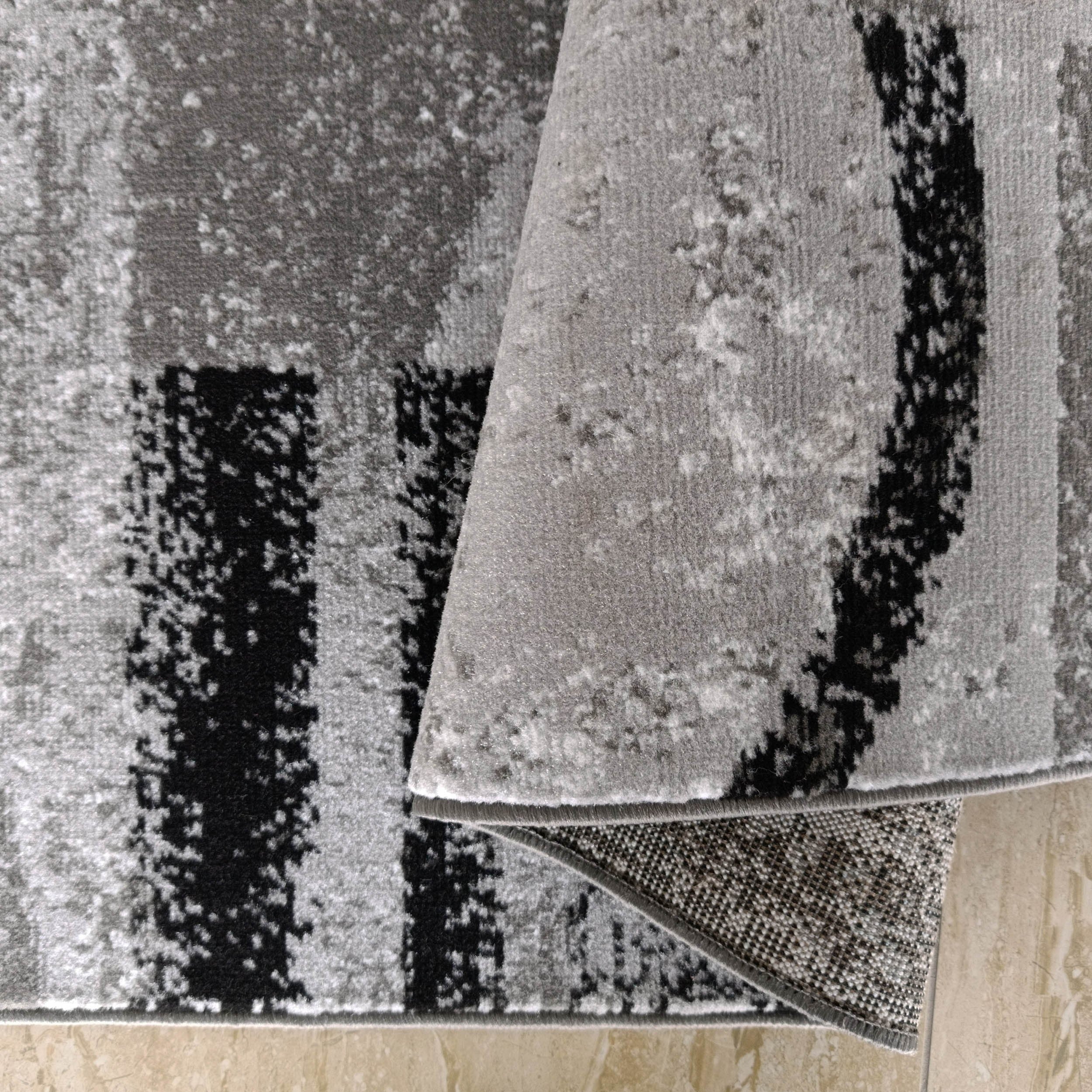 Tapis gris clair et noir à motif abstrait, partiellement roulé sur un carrelage beige, révélant son envers tissé