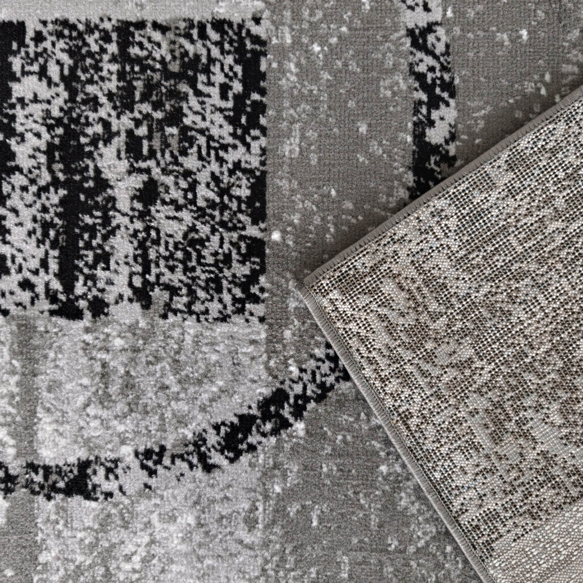 Tapis abstrait noir, gris clair et foncé avec un coin relevé dévoilant un envers gris à points noirs tissés