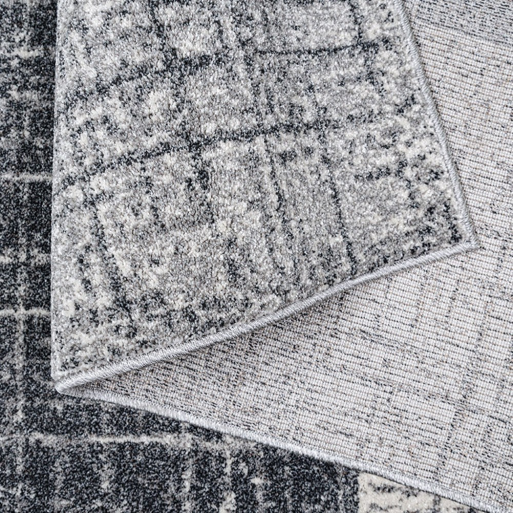 Tapis texturé gris et noir à motifs quadrillés abstraits, plié, dévoilant son envers tissé clair et un bord cousu