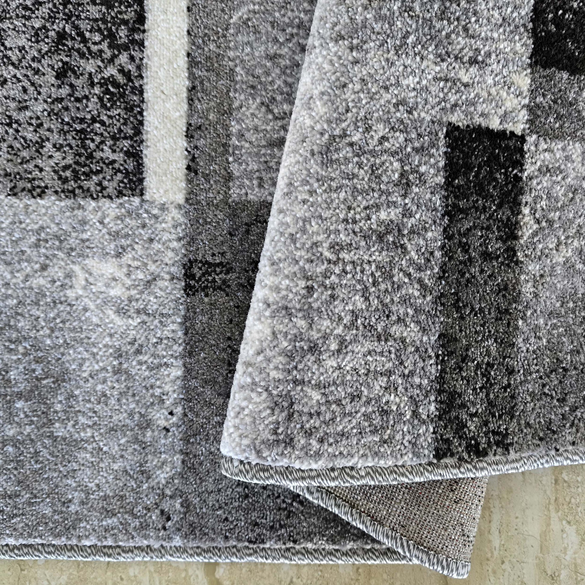 Tapis doux aux motifs géométriques noirs, gris et blancs, dont un coin est relevé montrant l'envers tissé