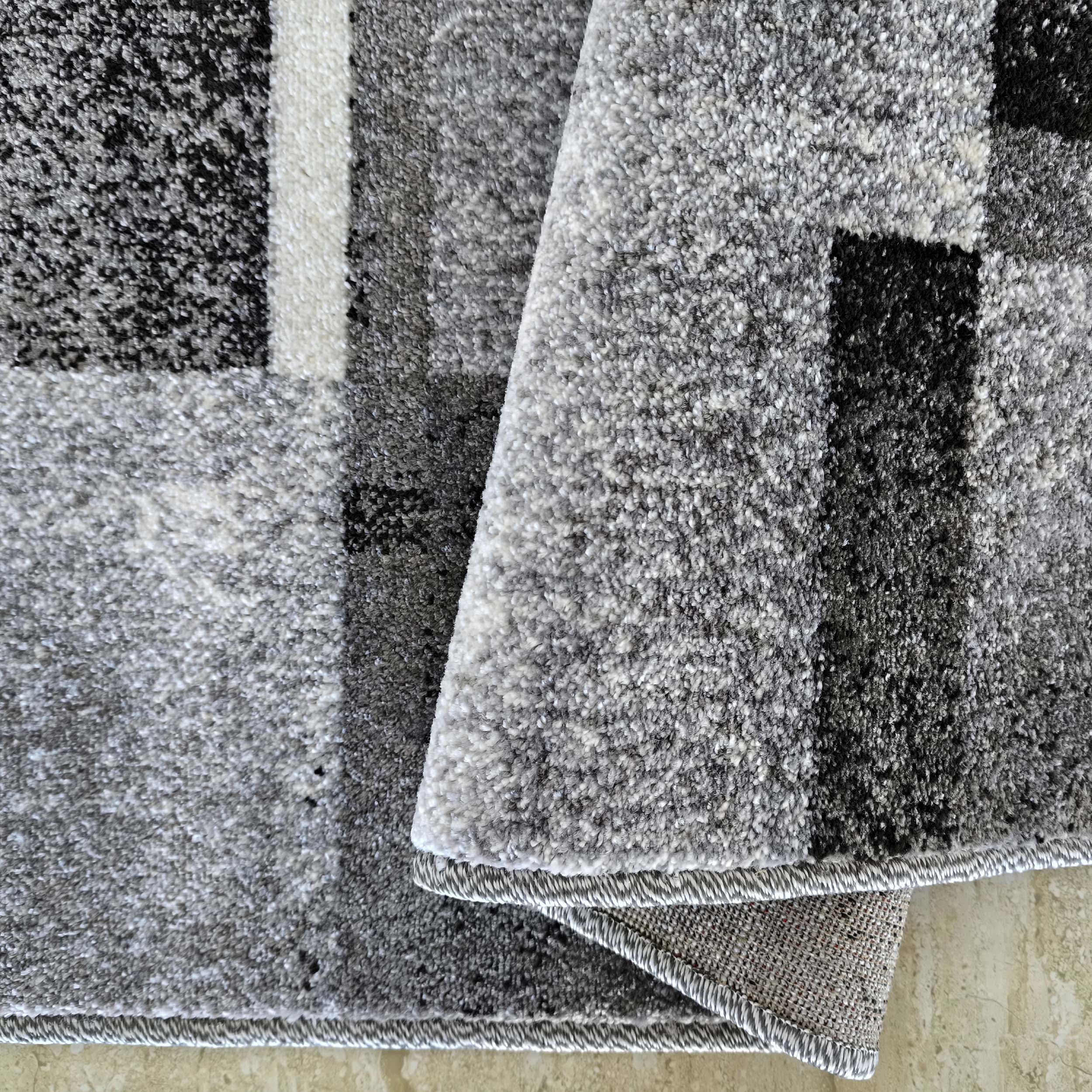 Tapis doux aux motifs géométriques noirs, gris et blancs, dont un coin est relevé montrant l'envers tissé