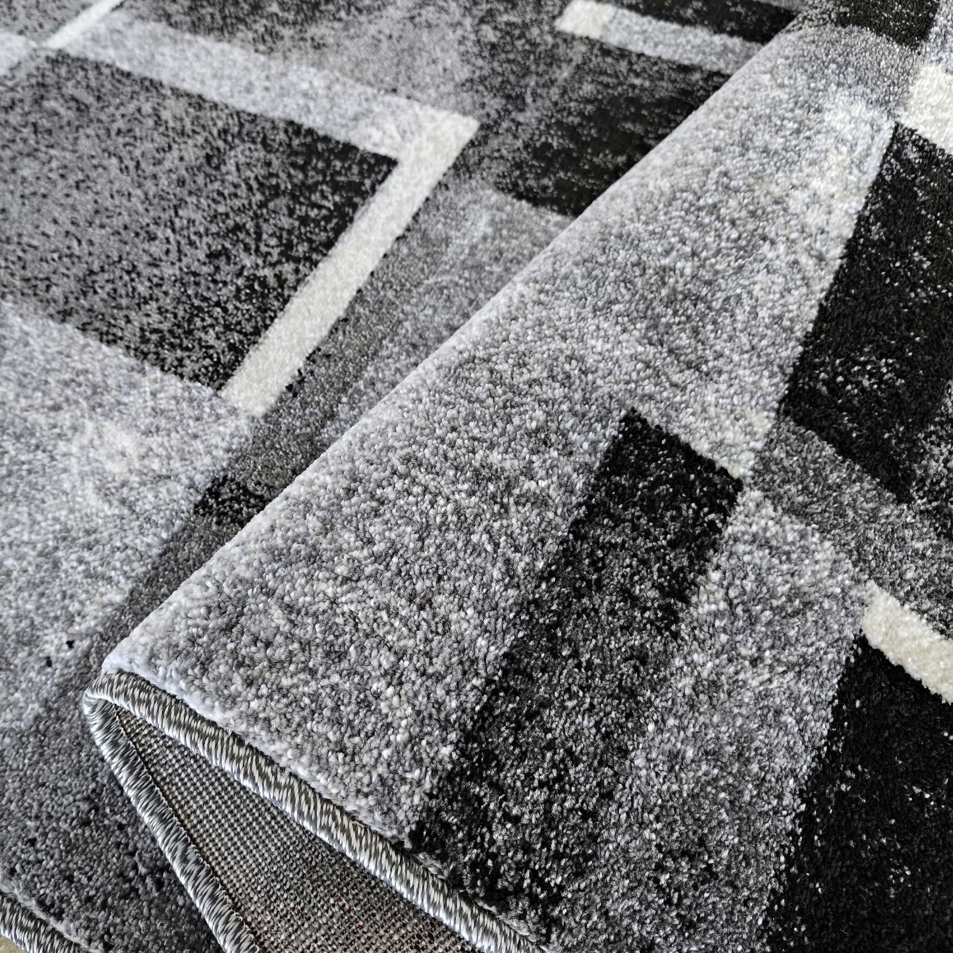 Tapis moelleux aux motifs géométriques gris, noirs et blancs, une partie est enroulée montrant le revers tissé
