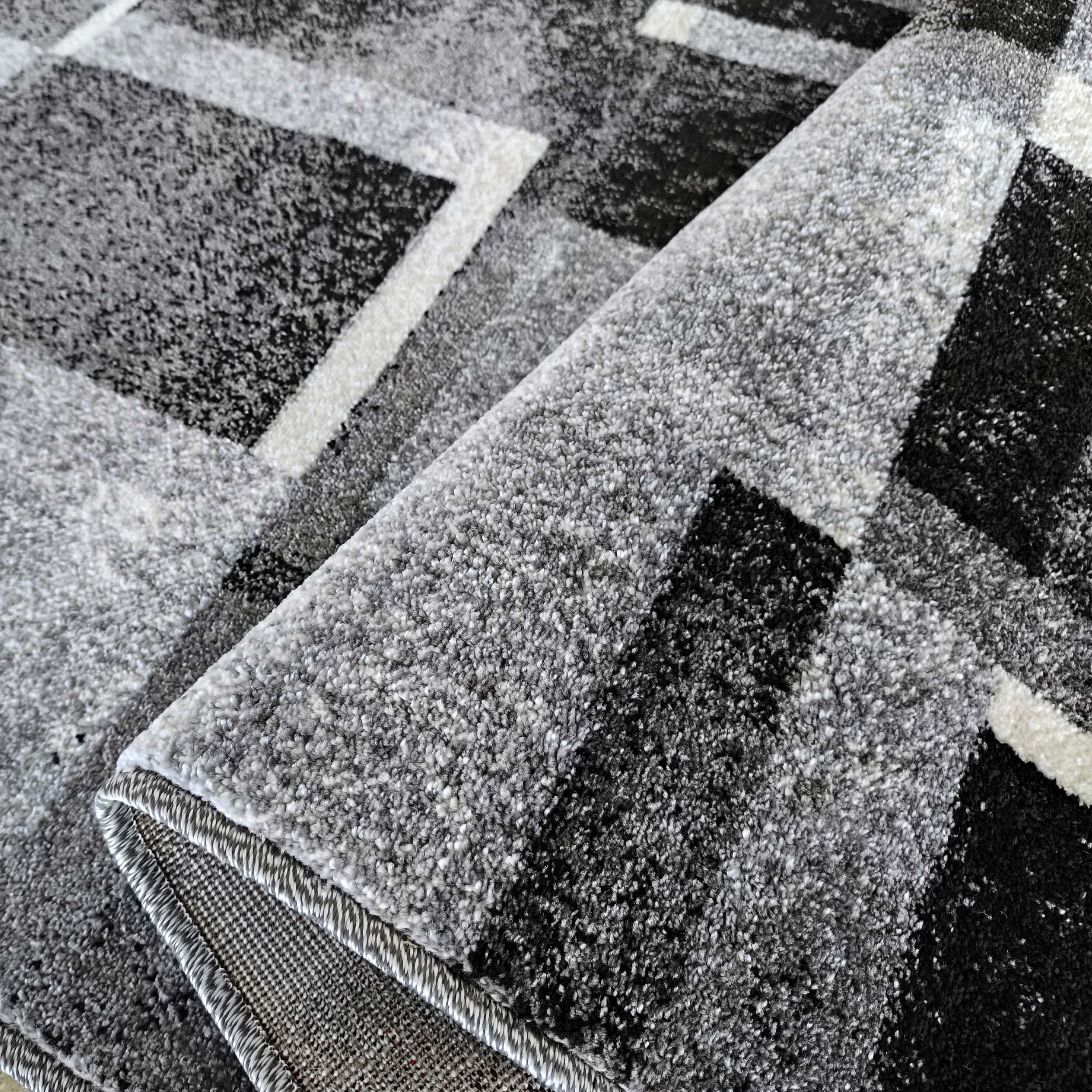 Tapis moelleux aux motifs géométriques gris, noirs et blancs, une partie est enroulée montrant le revers tissé