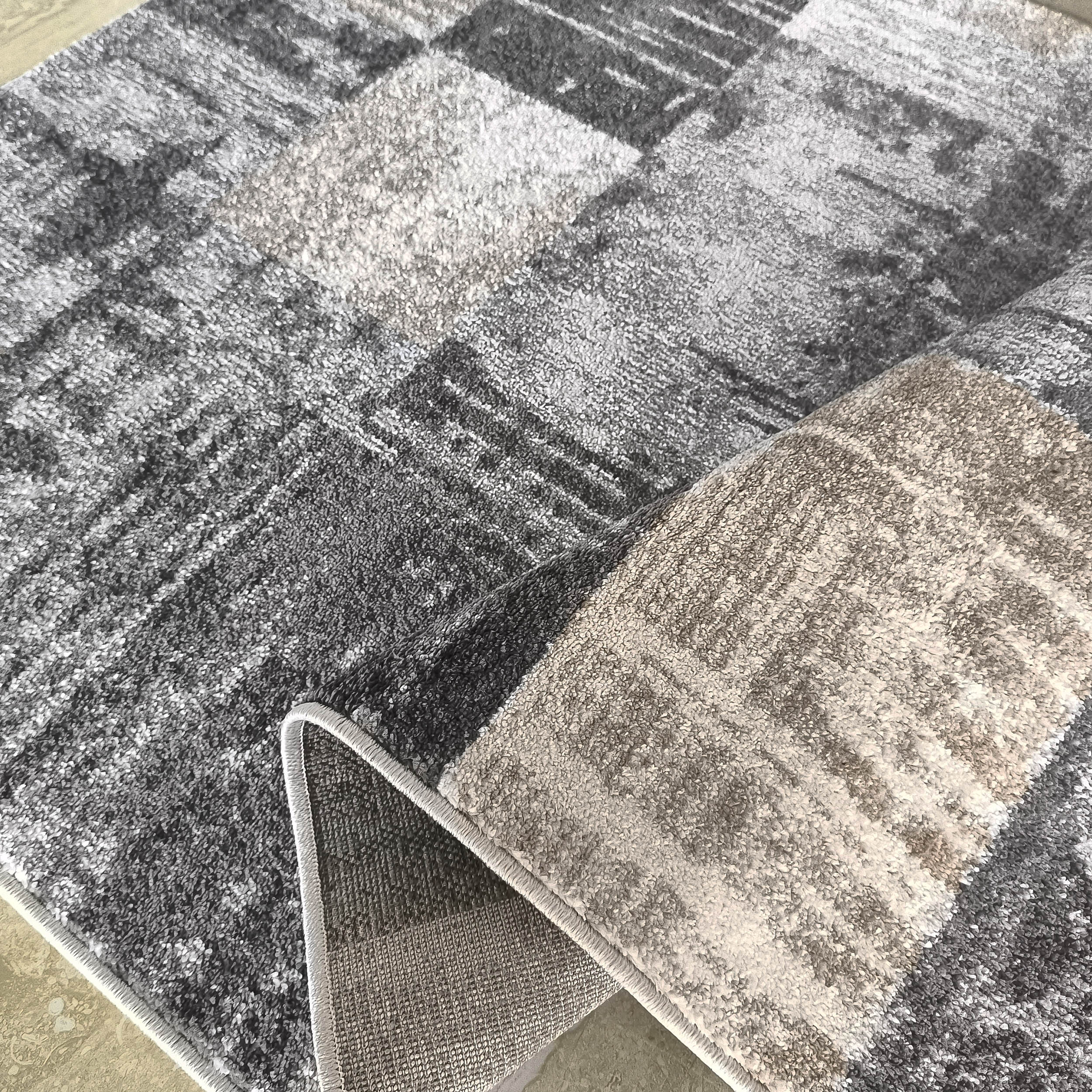 Tapis aux motifs abstraits de blocs gris, blancs et beiges, partiellement roulé révélant l'envers tissé