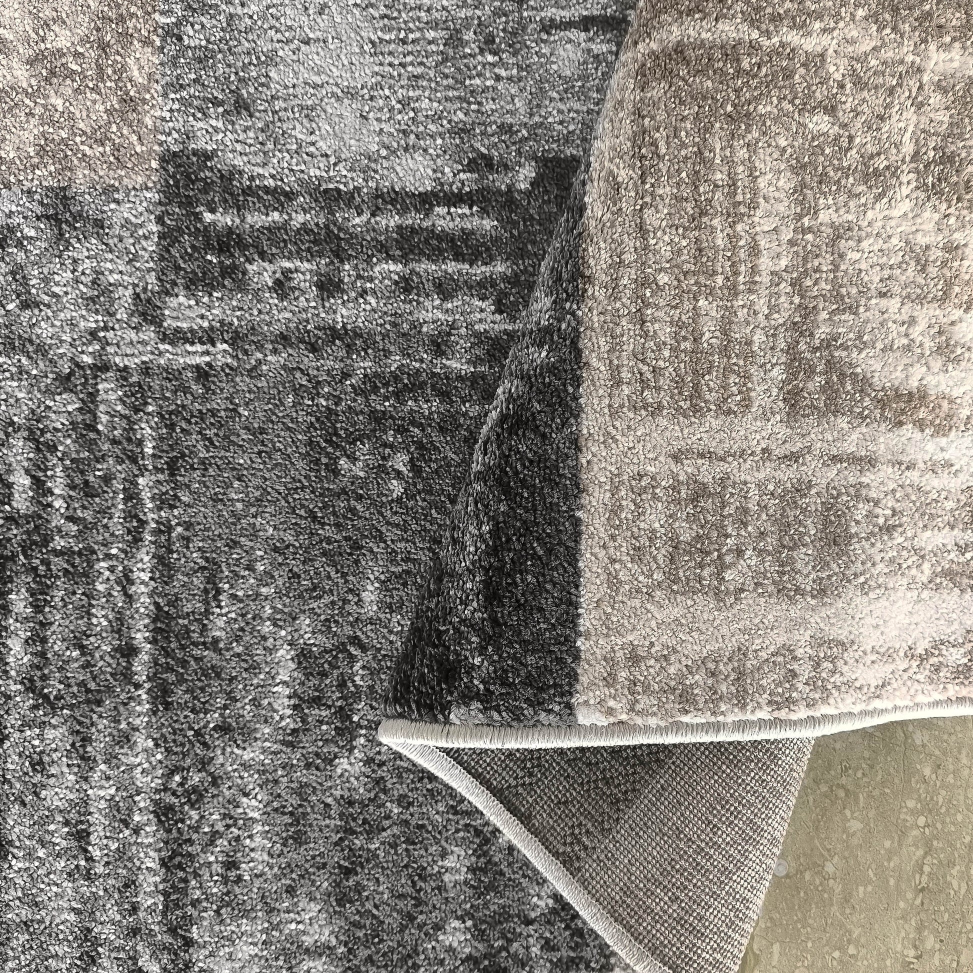 Tapis doux gris et beige à motifs abstraits, dont un bord est roulé pour montrer le dos et le sol