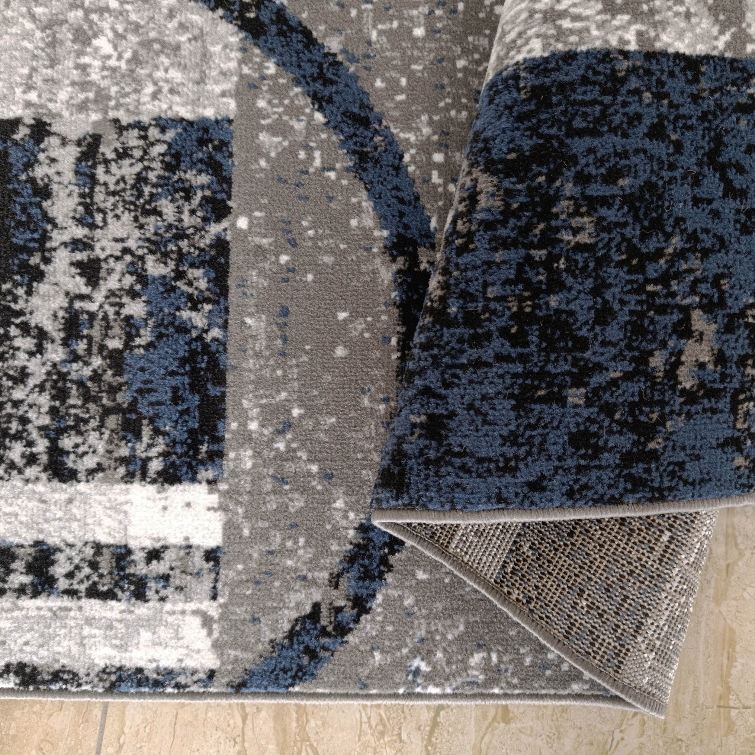 Tapis roulé à motif abstrait gris, bleu foncé, noir et blanc, son envers texturé visible sur un sol carrelé beige