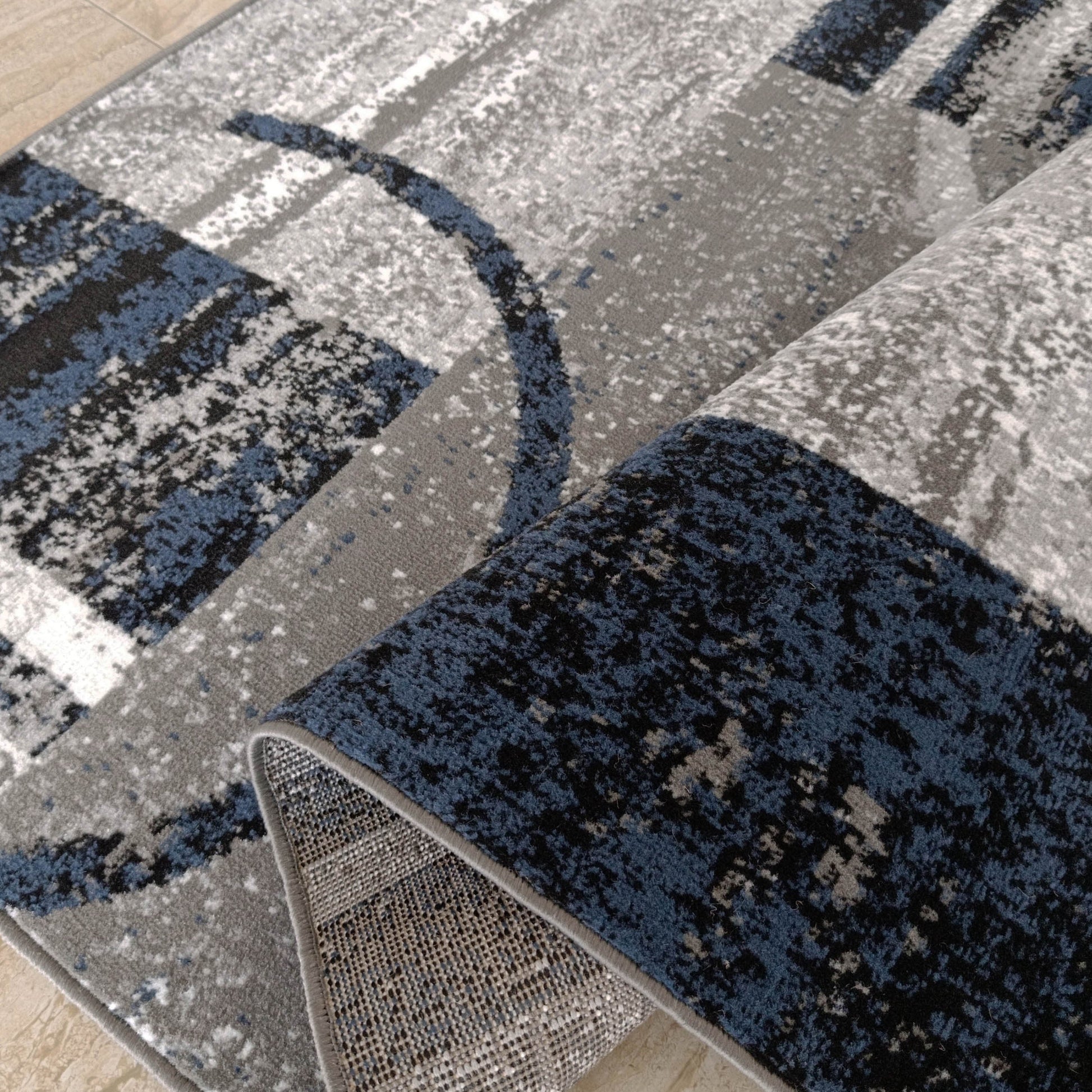 Tapis tissé abstrait gris, blanc, noir et bleu marine, partiellement roulé sur sol carrelé beige