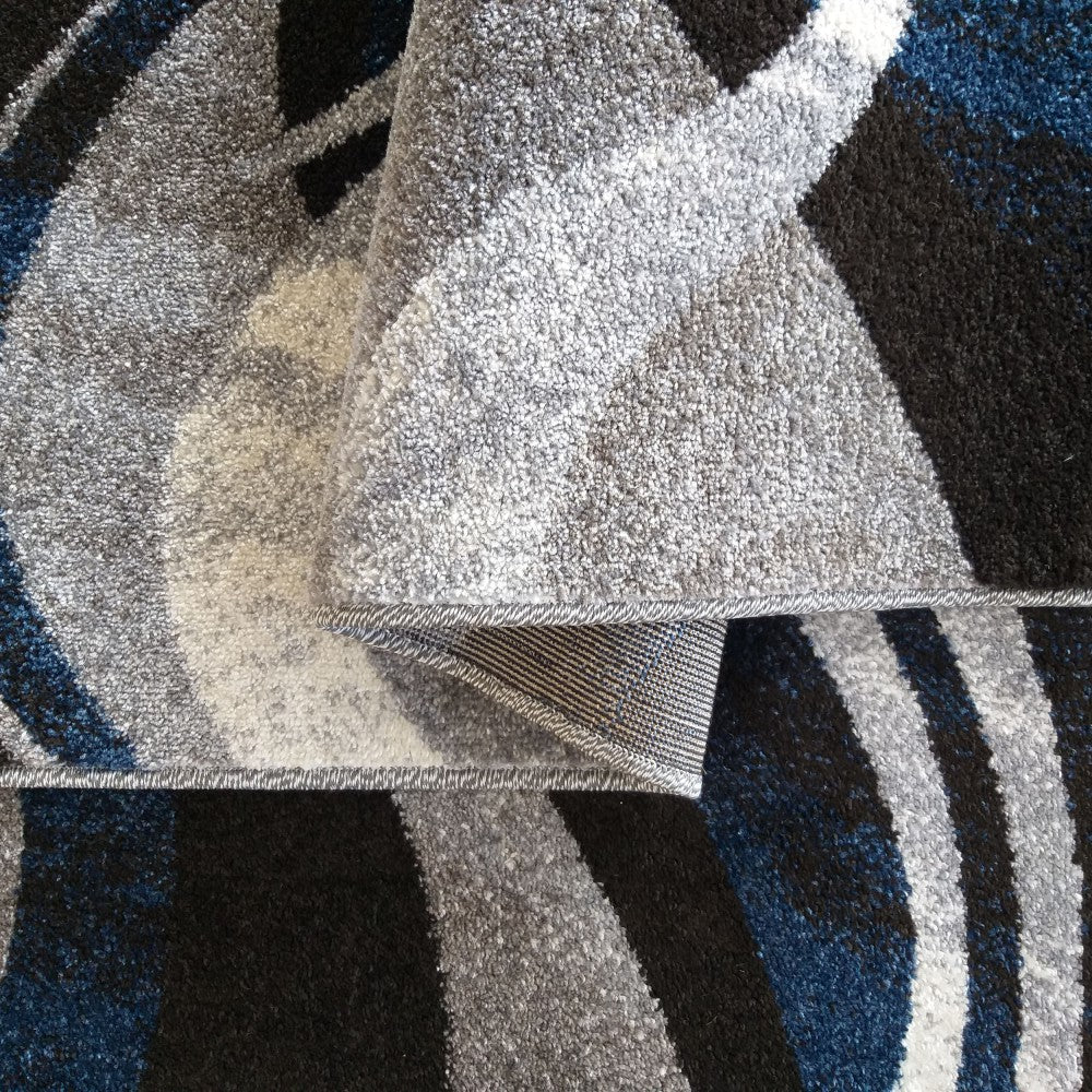Gros plan d'un tapis à poils courts. Il présente des motifs abstraits et ondulés en bleu, noir, gris et blanc, une partie étant pliée