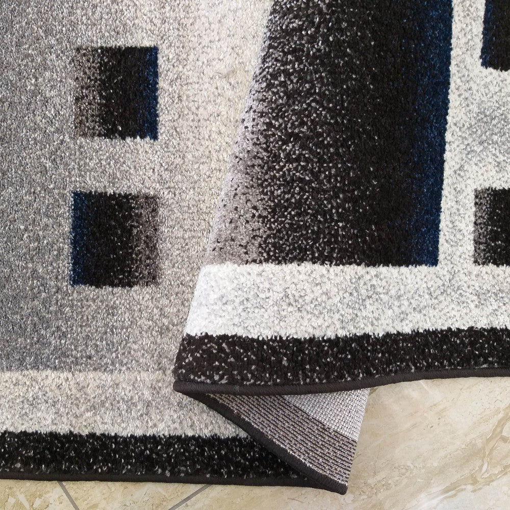 Tapis texturé gris, noir et blanc à motifs géométriques et dégradés, en partie roulé sur un carrelage beige