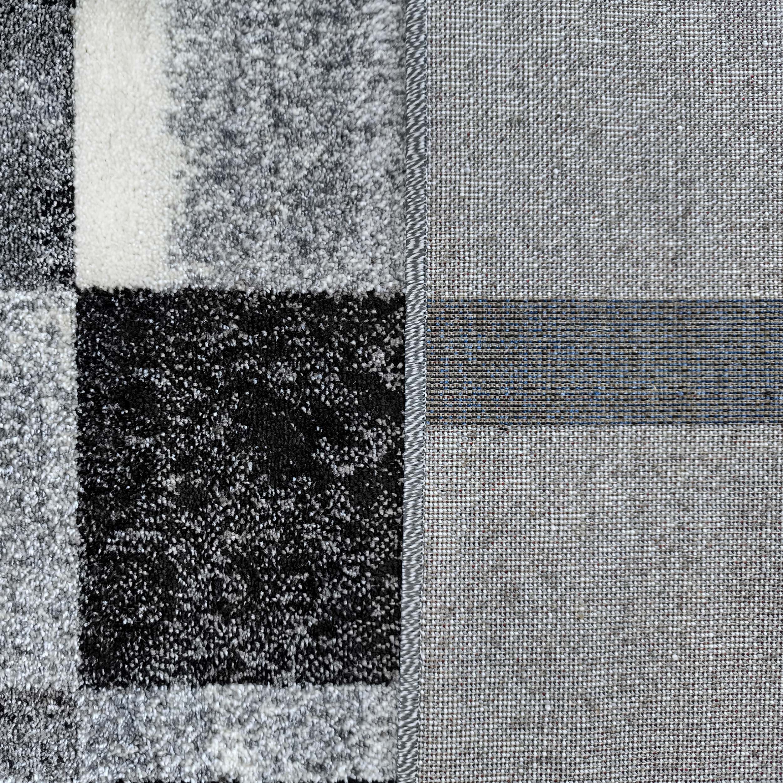 Tapis à motif de grands carrés noirs, blancs et gris sur le dessus texturé, montrant aussi le revers tissé gris avec des fils bleus