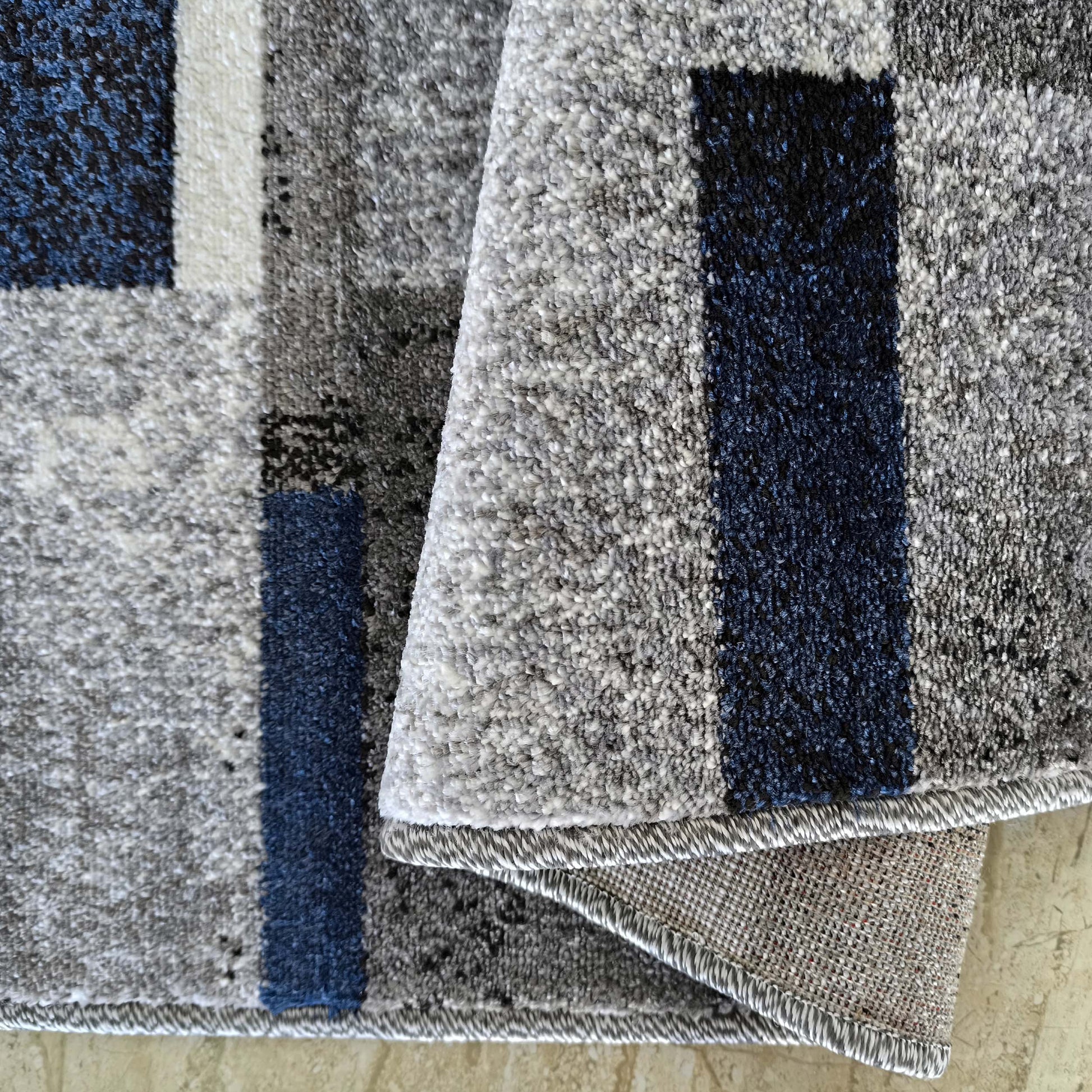 Tapis contemporain aux blocs de couleur gris clair, foncé, bleu et blanc, avec une texture bouclée et son revers plié