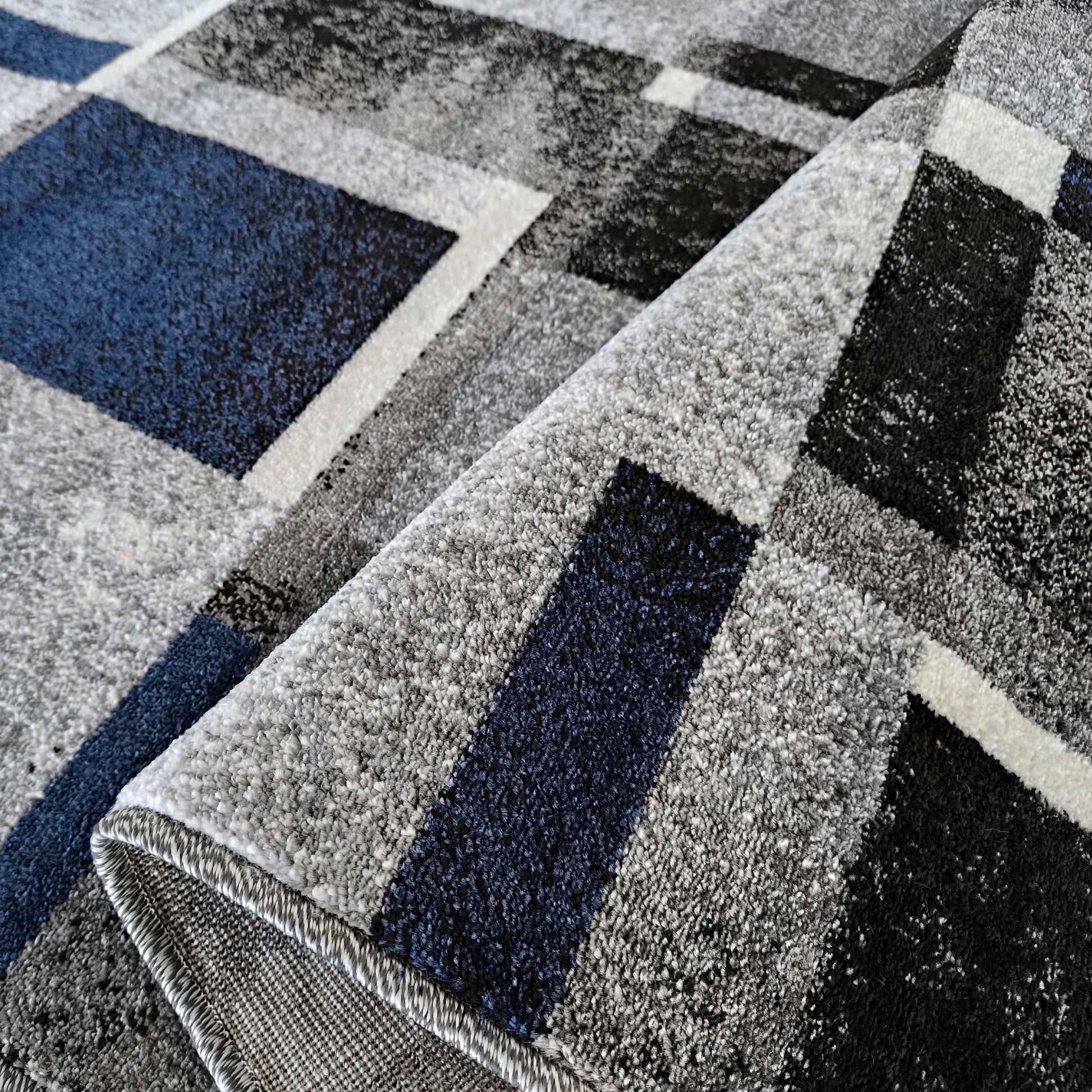 Tapis à motifs géométriques de blocs bleus, gris, blancs et noirs. Sa texture en poils est visible et une partie est enroulée