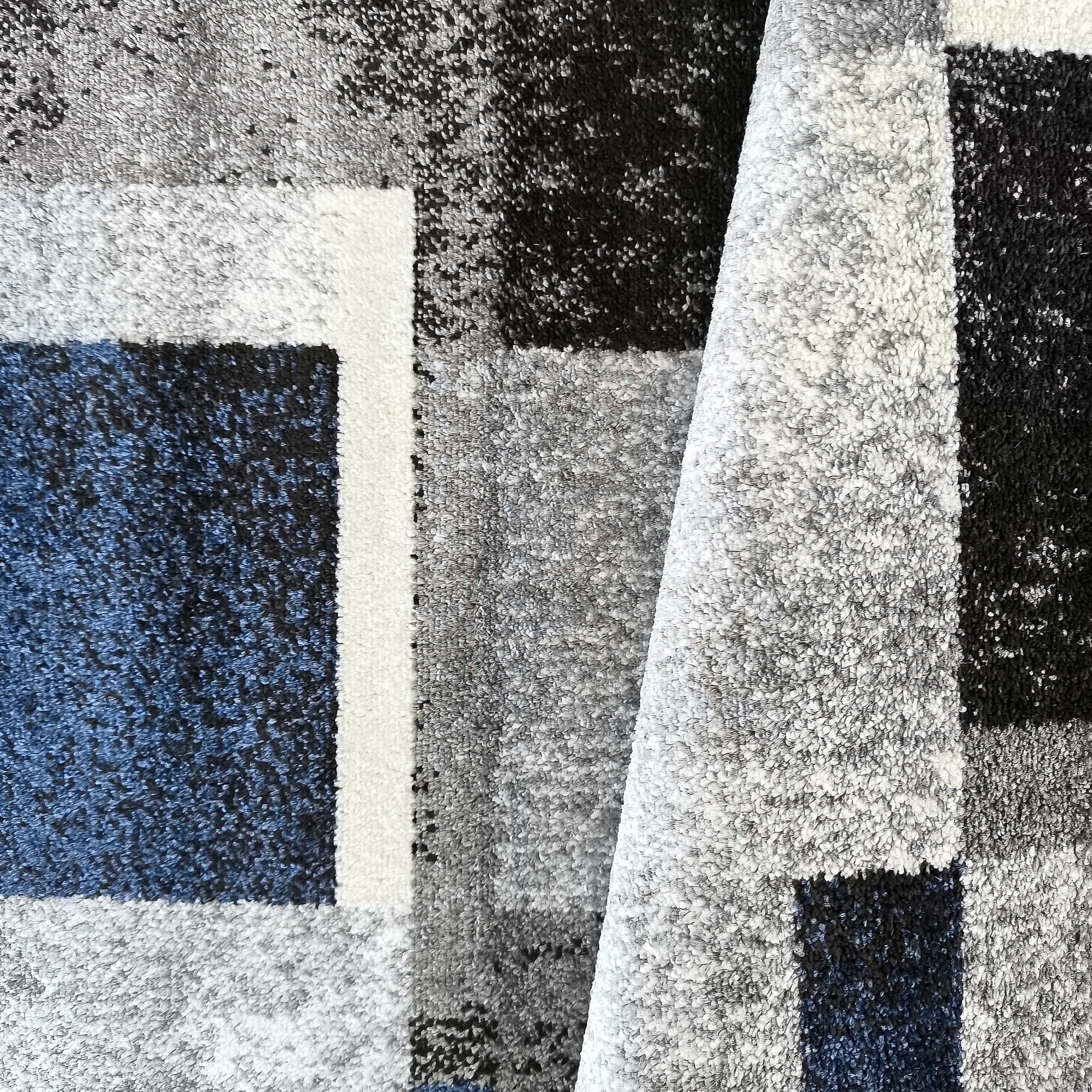 Tapis à poils longs aux motifs géométriques gris, noir, blanc et bleu foncé, avec une partie légèrement repliée