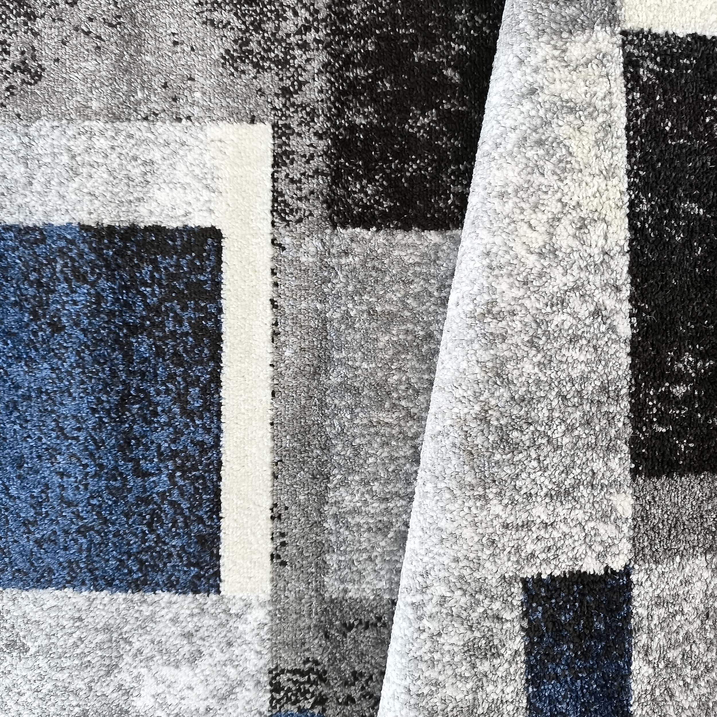 Tapis à poils longs aux motifs géométriques gris, noir, blanc et bleu foncé, avec une partie légèrement repliée