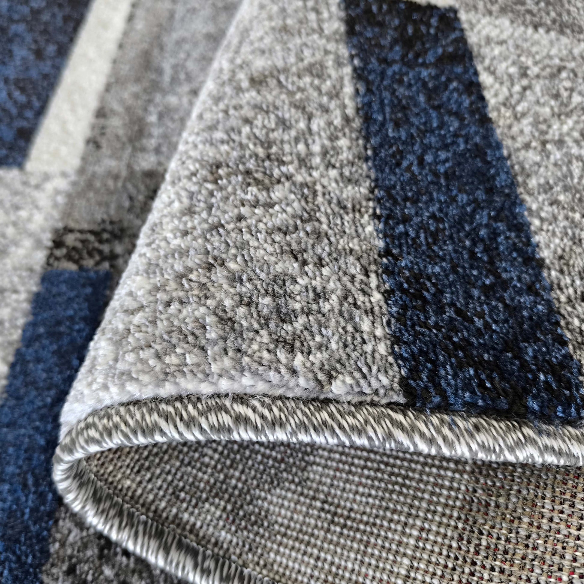 Gros plan d'un tapis aux motifs géométriques gris, bleu foncé et blanc, sa surface texturée et son dessous tissé visibles