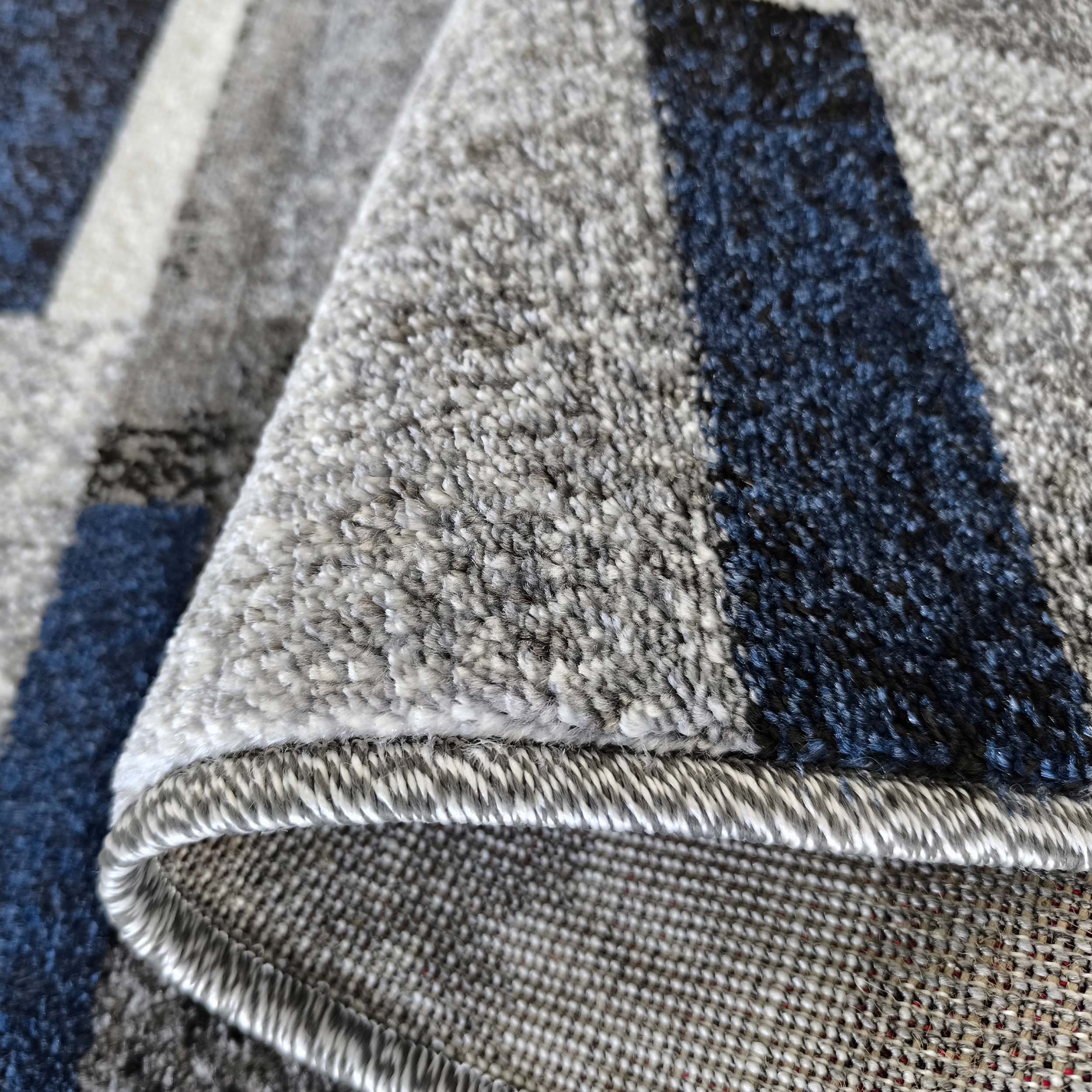 Gros plan d'un tapis aux motifs géométriques gris, bleu foncé et blanc, sa surface texturée et son dessous tissé visibles