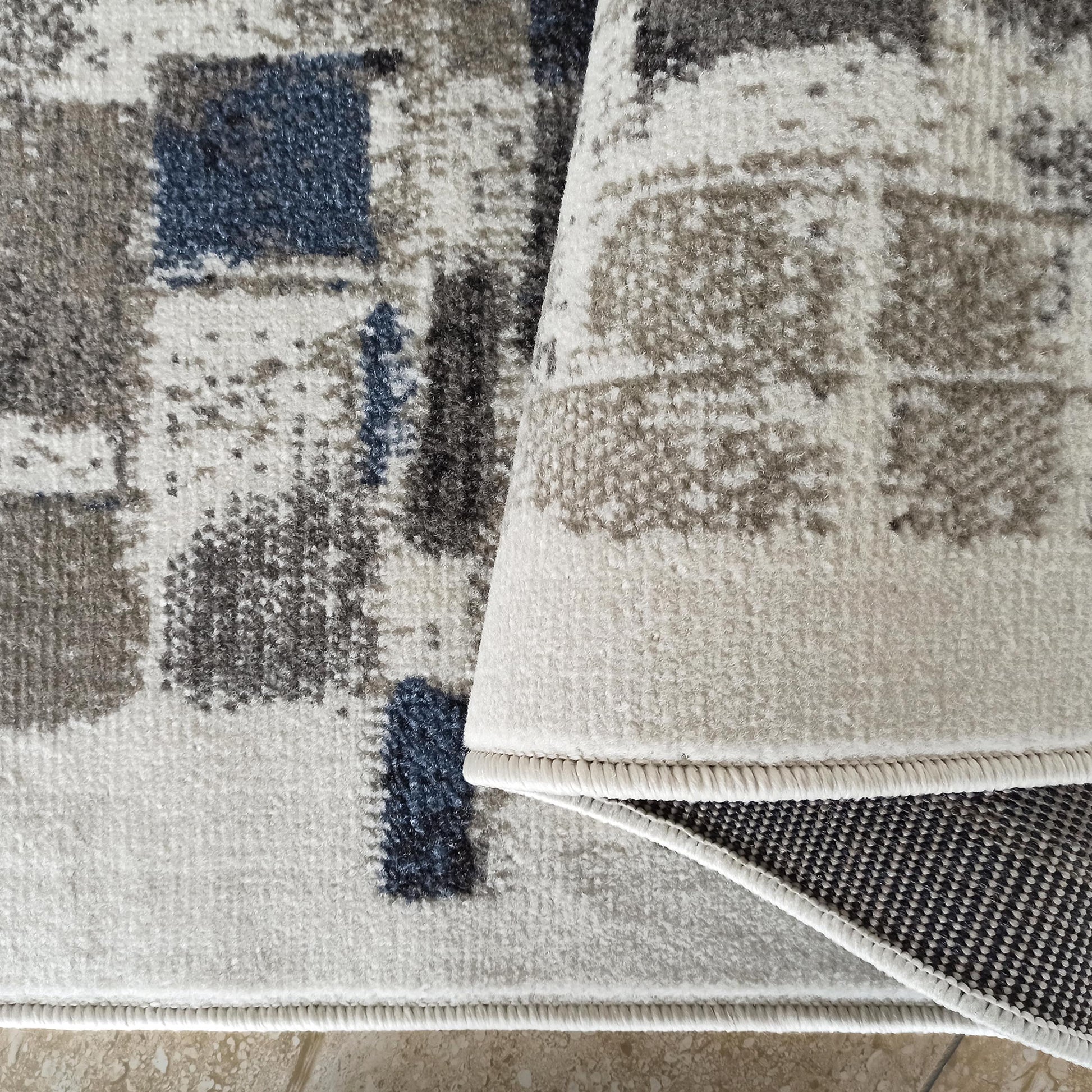 Tapis à poils courts aux motifs abstraits dans les tons gris, crème et bleu. Une partie est roulée, dévoilant son envers tissé